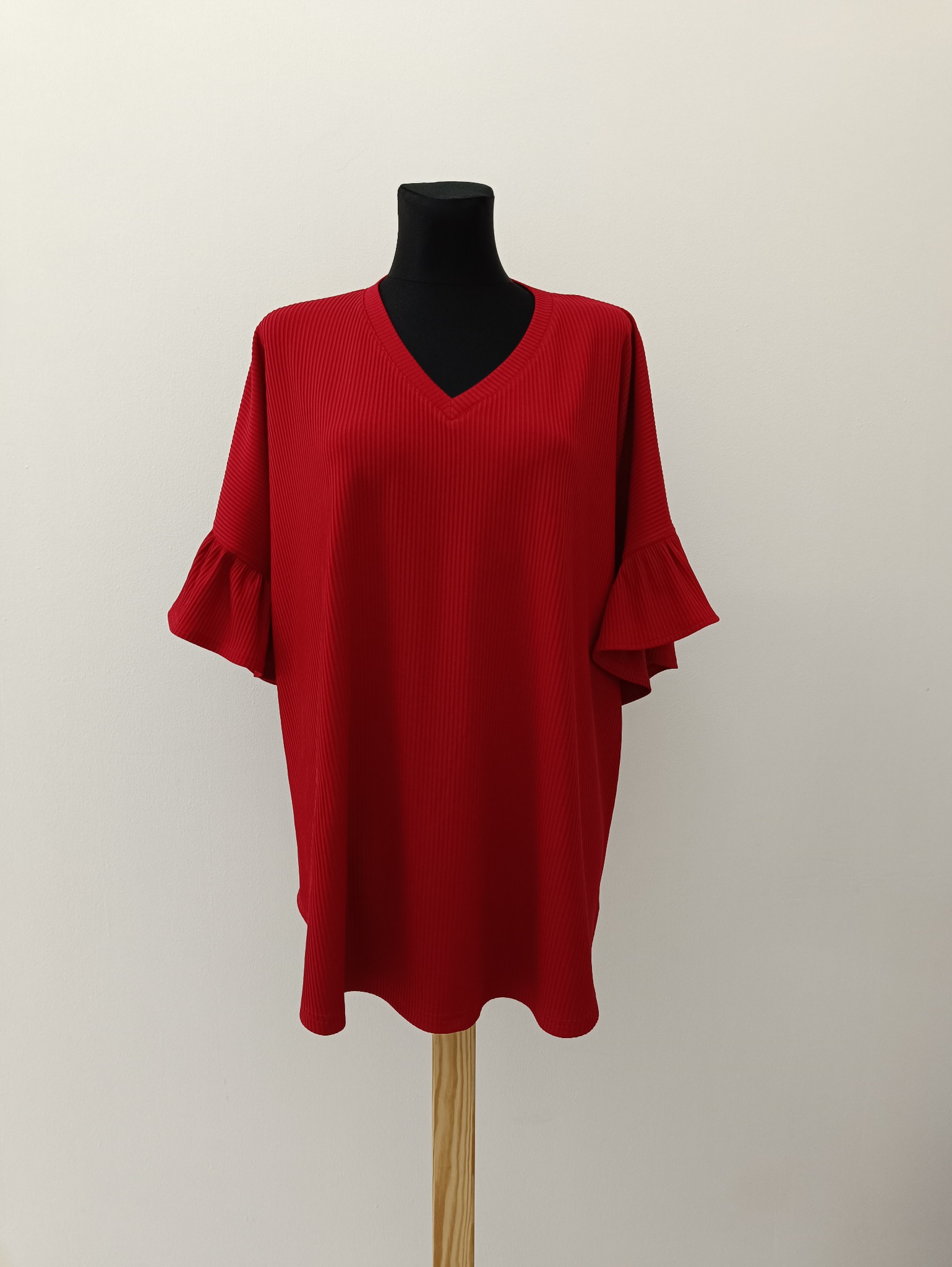 Czerwona, prążkowana bluzka z krótkimi, falowanymi rękawami na czarnym manekinie. Tło stanowi kremowa ściana, a całość prezentuje się schludnie i profesjonalnie.