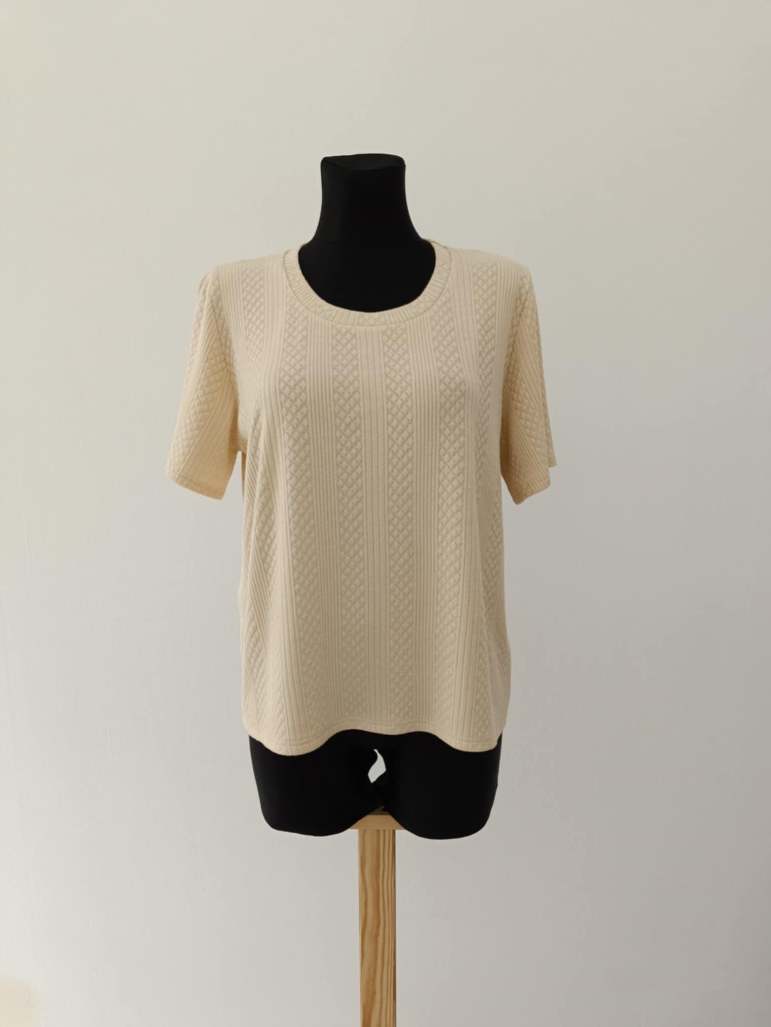 Beżowa bluzka z krótkim rękawem o fakturze w romby, prezentowana na czarnym manekinie na drewnianej podstawie, na tle jasnej ściany.