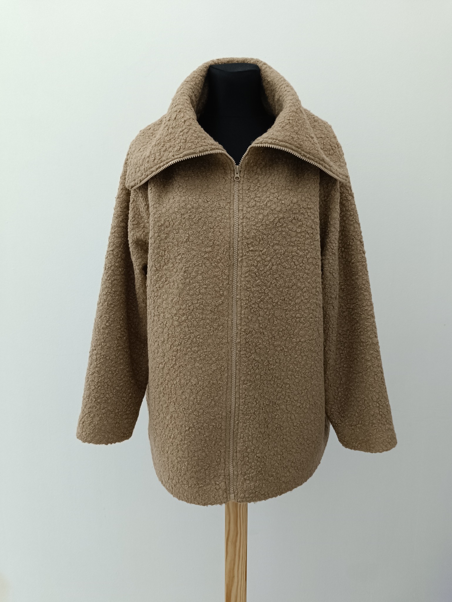 Beżowa, zapinana na zamek kurtka z efektem baranka, na czarnym manekinie. Widoczna faktura materiału i szeroki kołnierz. Neutralne tło, drewniana podstawa manekina.