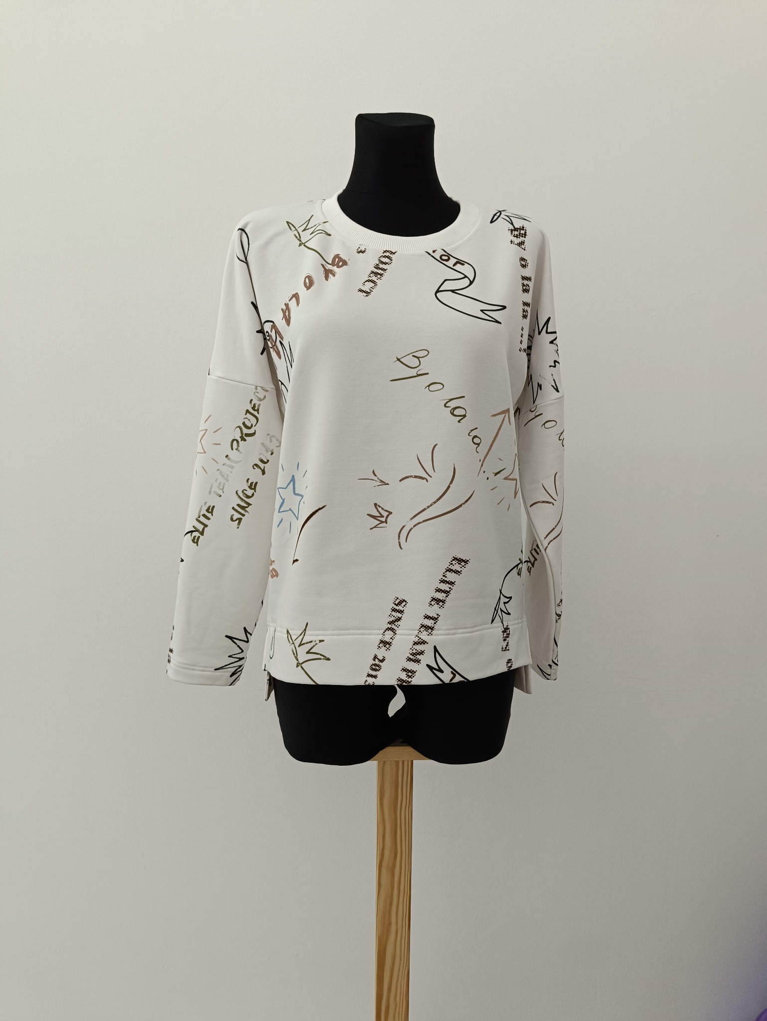 Biała bluza z długim rękawem i nadrukami, prezentowana na czarnym manekinie na drewnianej podstawie. Minimalistyczne tło, oświetlenie studyjne. Widoczne detale nadruku.