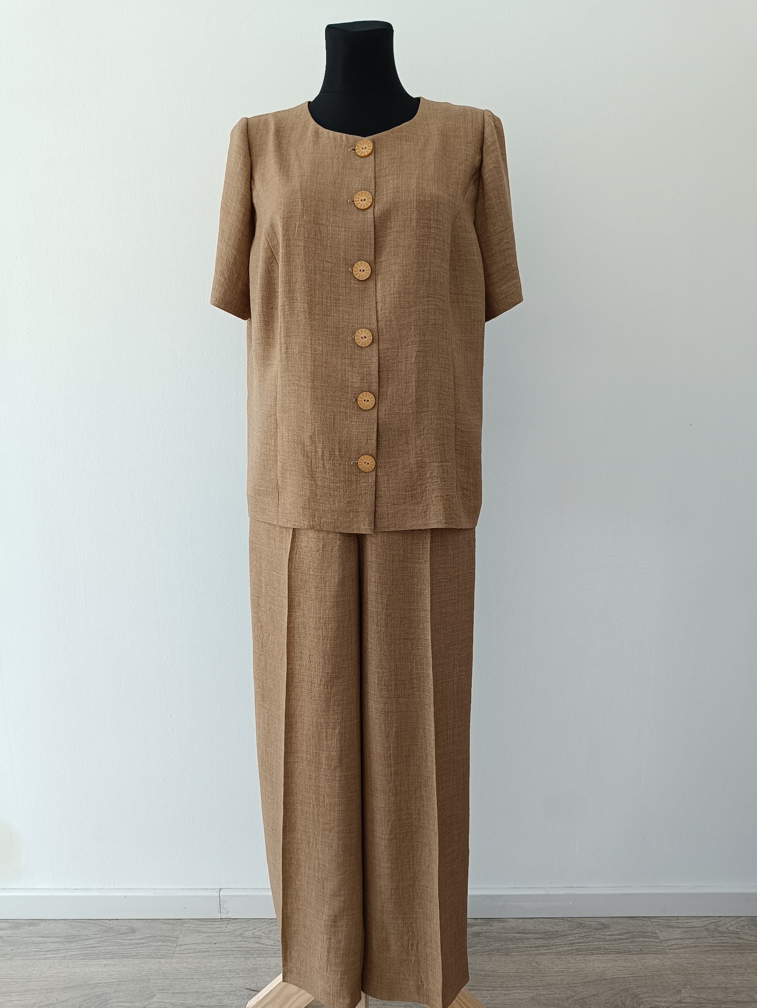 Brązowy, damski komplet odzieżowy z krótkim rękawem, zapinany na drewniane guziki, prezentowany na manekinie na tle białej ściany i drewnianej podłogi.