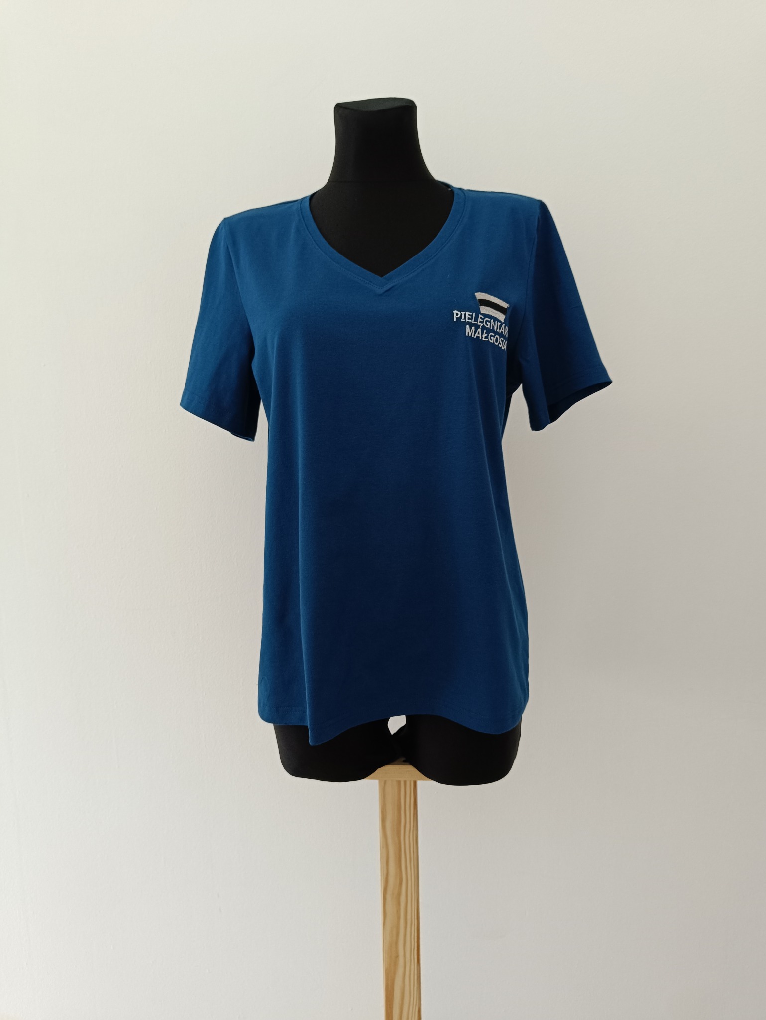 Niebieska koszulka z krótkim rękawem i haftowanym napisem na manekinie na białym tle. Prosty krój, dekolt w serek. Widoczny detal haftu.