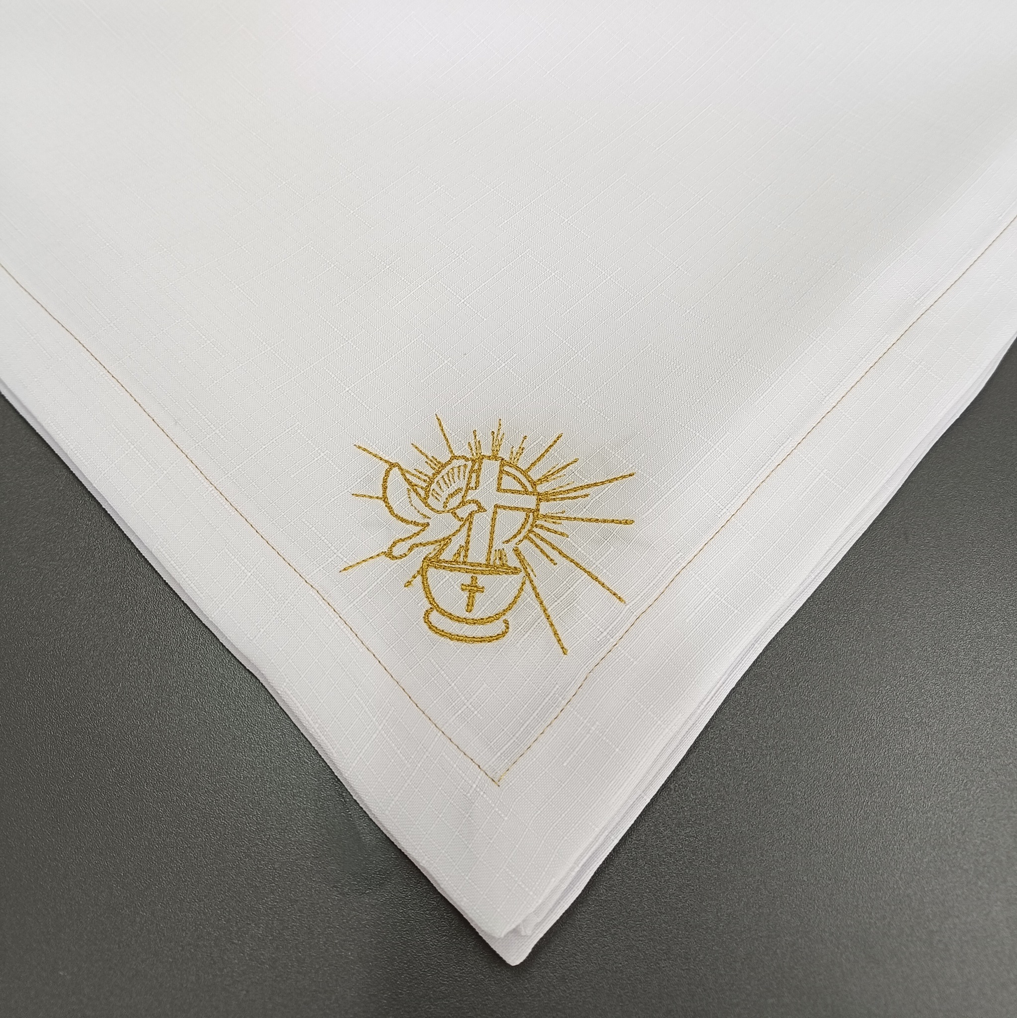 Biała, lniana serwetka komunijna z haftowanym kielichem, krzyżem i gołębicą w złotym kolorze. Starannie wykończona, leży na szarym tle.
