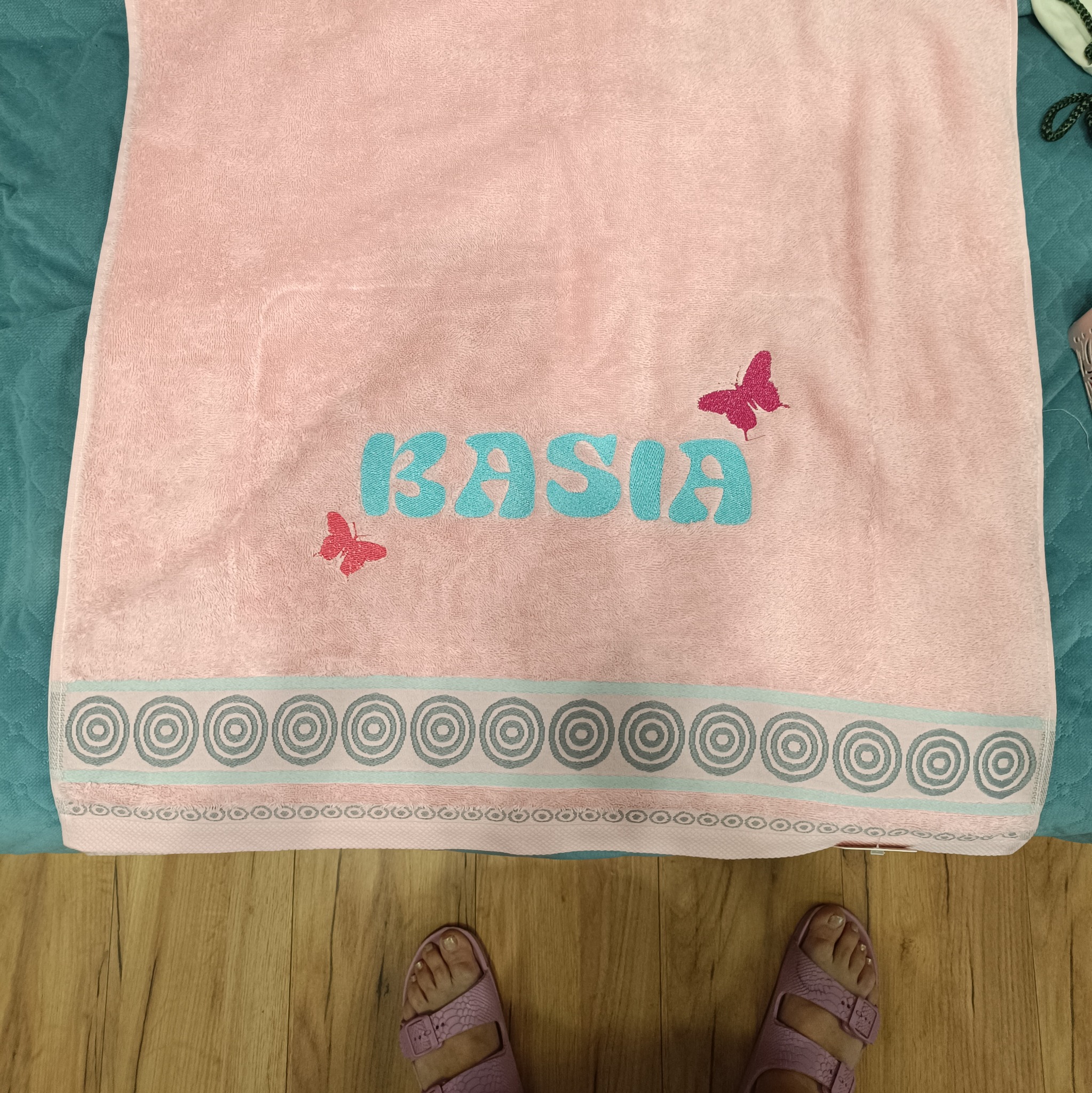 Różowy ręcznik z imieniem 'Kasia' i haftowanymi motylami, leżący na łóżku. Widoczne stopy w fioletowych sandałach na drewnianej podłodze.