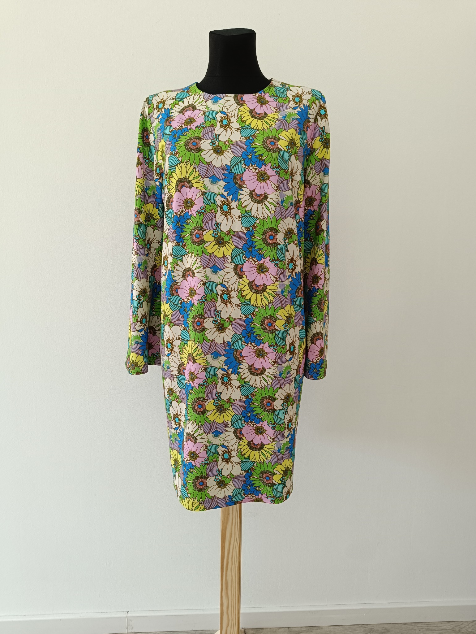 Sukienka mini z długim rękawem, w kwiatowy wzór w stylu retro, prezentowana na czarnym manekinie na tle białej ściany. Widoczna faktura materiału i detale szycia.