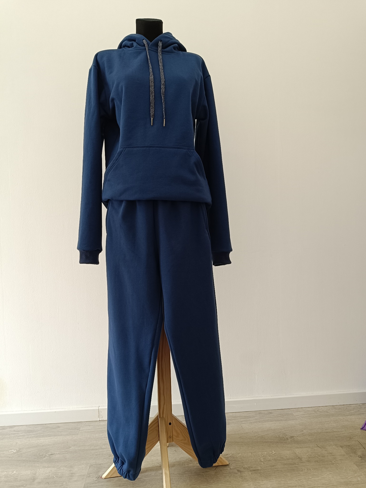 Granatowy dres damski z kapturem, prezentowany na manekinie na tle białej ściany. Bluza z kieszenią typu kangurka, spodnie dresowe ze ściągaczami.