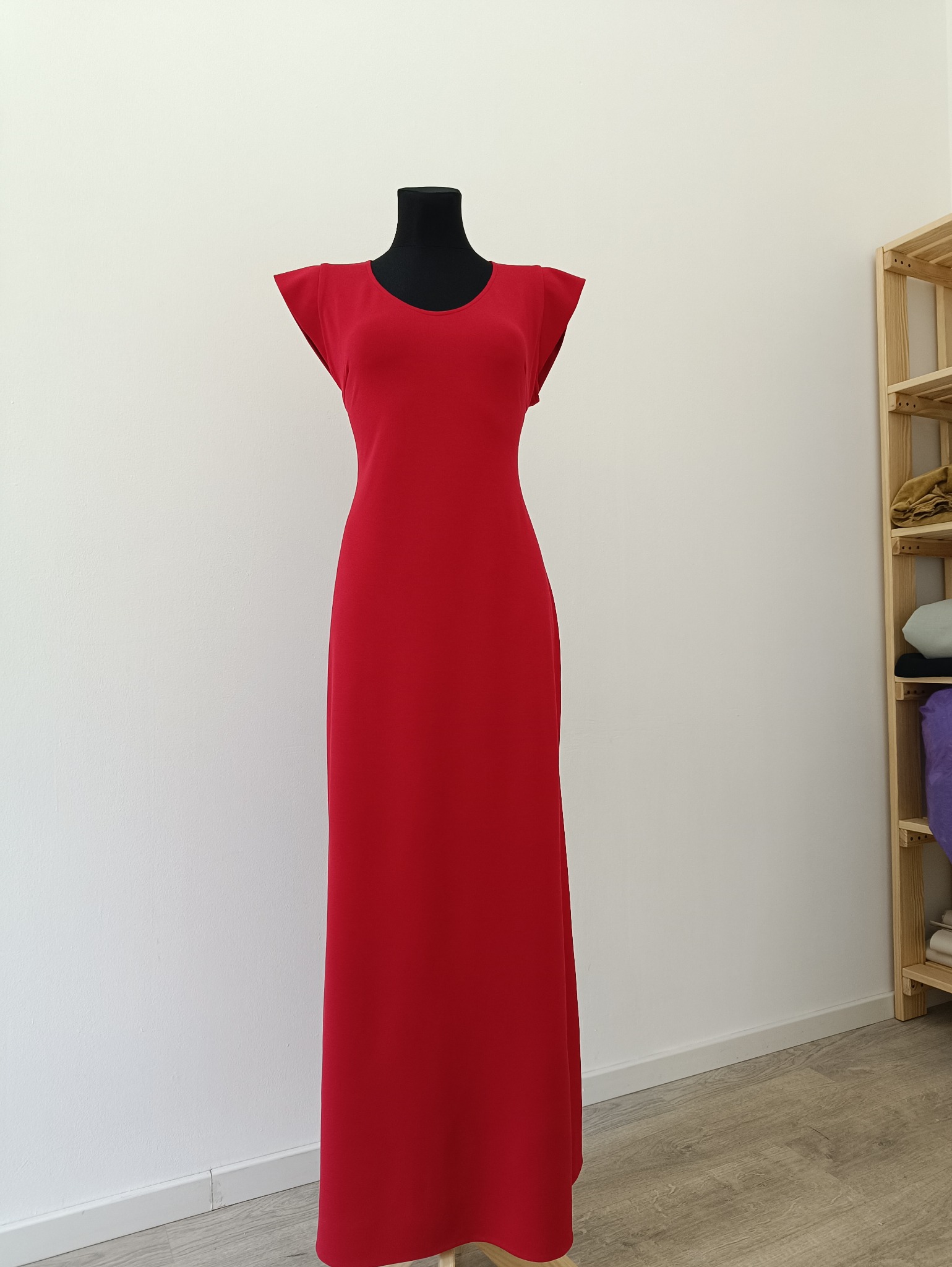 Czerwona, długa suknia z krótkim rękawem, zaprezentowana na czarnym manekinie na tle białej ściany i drewnianego regału. Minimalistyczny design.