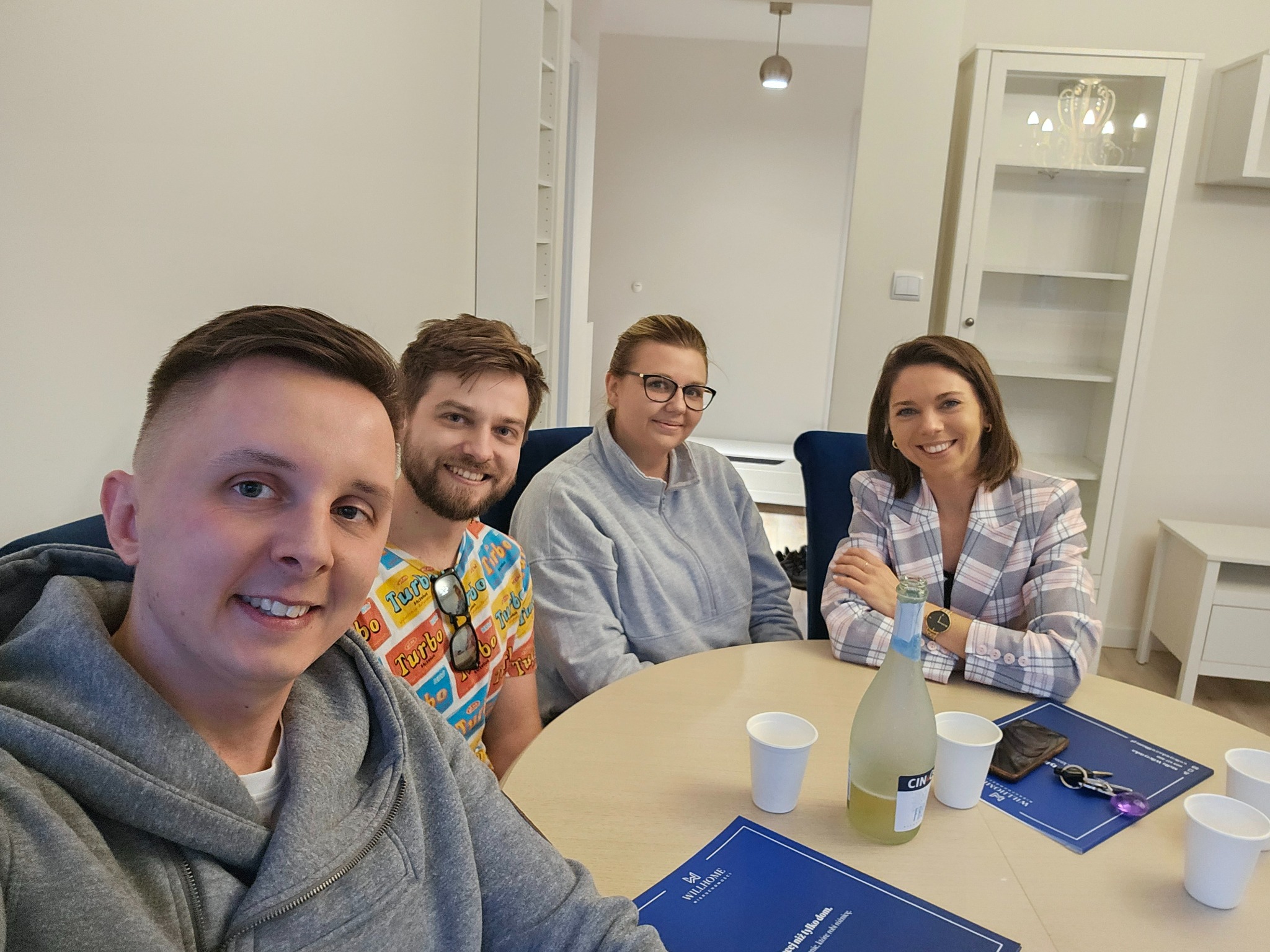 Grupa czterech uśmiechniętych osób siedzi przy okrągłym stole w jasnym pomieszczeniu; na stole butelka napoju, kubki, klucze i materiały informacyjne z logo Willhome nieruchomości.