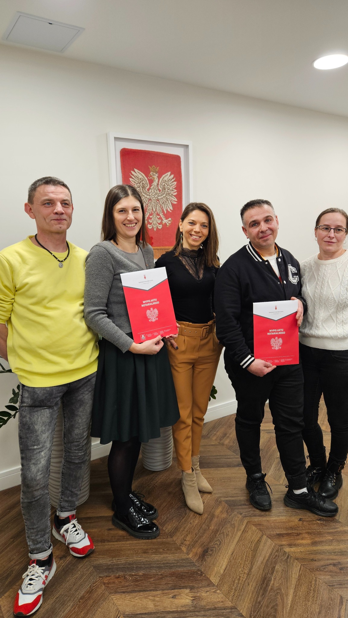 Grupa osób trzymająca czerwone dokumenty 'Wypis Aktu Notarialnego' z godłem Polski, stojąca na tle ściany z obrazem orła w koronie.
