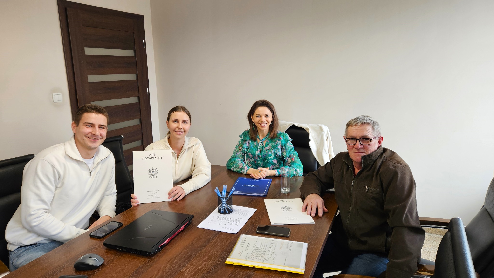 Cztery osoby siedzą przy stole w biurze, dwie z nich trzymają dokument 'Akt Notarialny', na stole leżą dokumenty, długopisy i laptop.