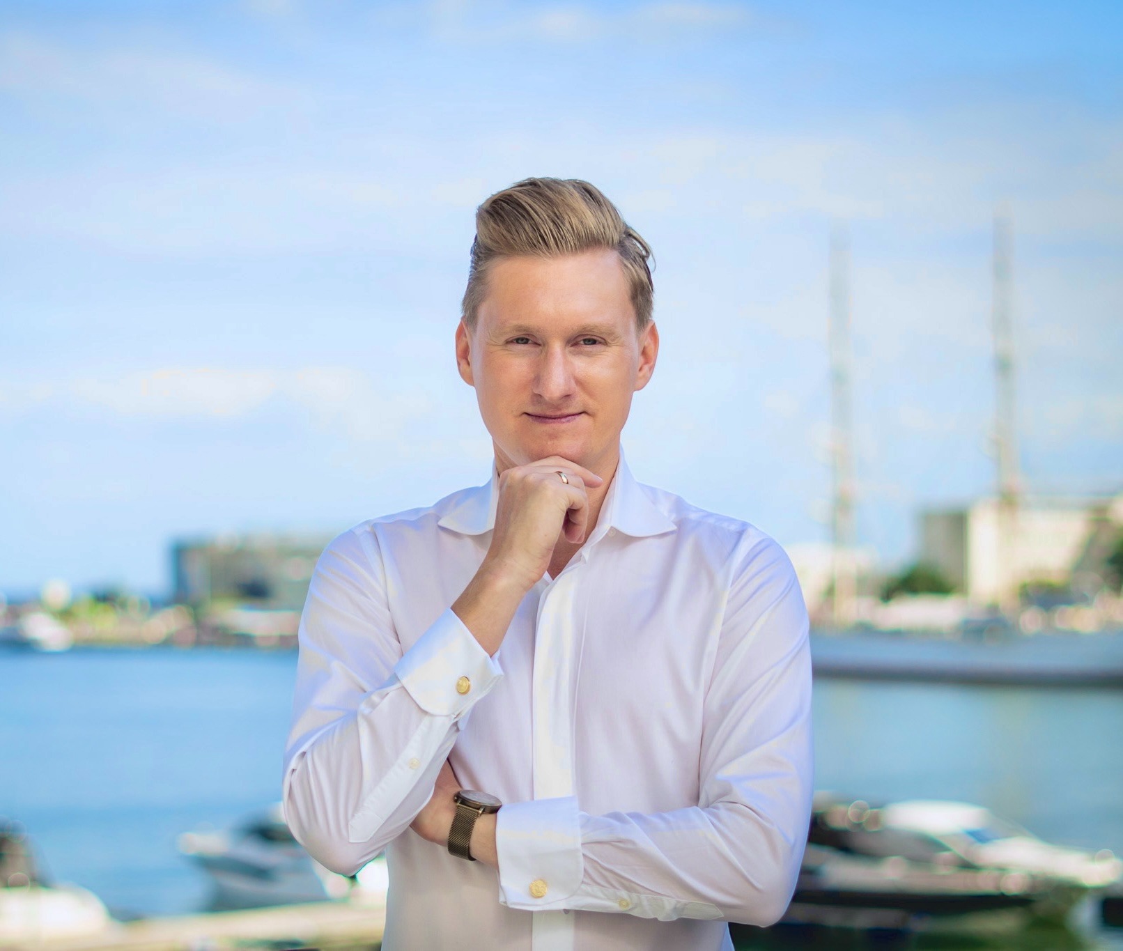 Portret mężczyzny w białej koszuli z podwiniętymi rękawami i złotymi spinkami, z zegarkiem na ręce, zamyślony, z ręką pod brodą na tle portu z jachtami i błękitnego nieba.