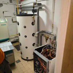 Instalacja w kotłowni: biały zasobnik ciepłej wody, rozbudowana instalacja rur, otwarty panel pompy ciepła z widocznymi miedzianymi rurkami i okablowaniem, obok widoczny zielony panel sterowania...