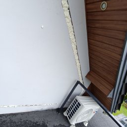 STACH Wojciech Skoneczny - Biała jednostka zewnętrzna klimatyzacji zamontowana na płaskim dachu z widocznym ociepleniem ściany pianką montażową i fragmentem drewnianej podbitki dachowej.