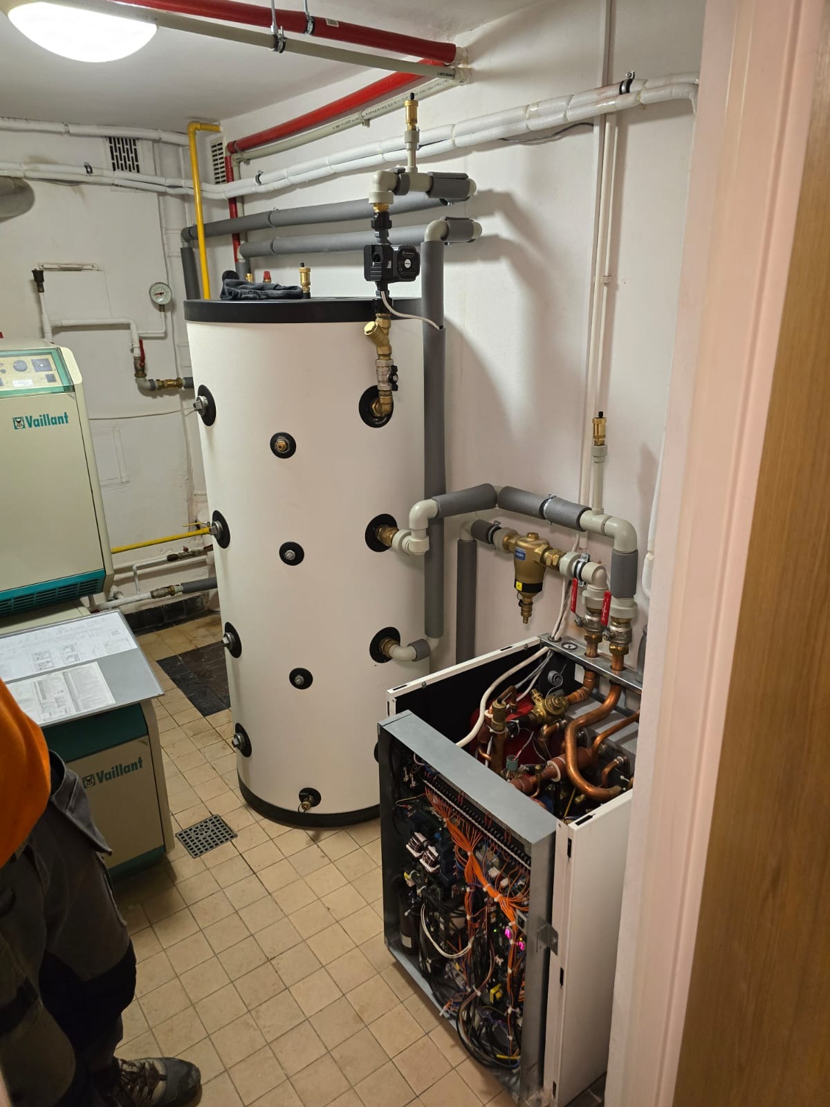 Instalacja w kotłowni: biały zasobnik ciepłej wody, rozbudowana instalacja rur, otwarty panel pompy ciepła z widocznymi miedzianymi rurkami i okablowaniem, obok widoczny zielony panel sterowania...