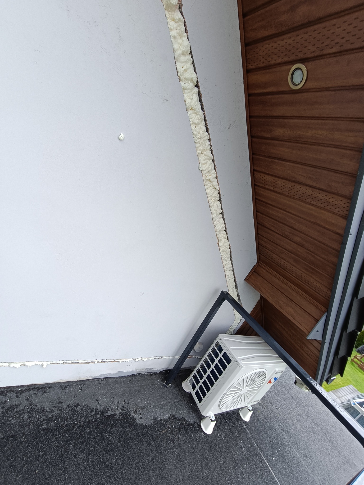 Biała jednostka zewnętrzna klimatyzacji zamontowana na płaskim dachu z widocznym ociepleniem ściany pianką montażową i fragmentem drewnianej podbitki dachowej.