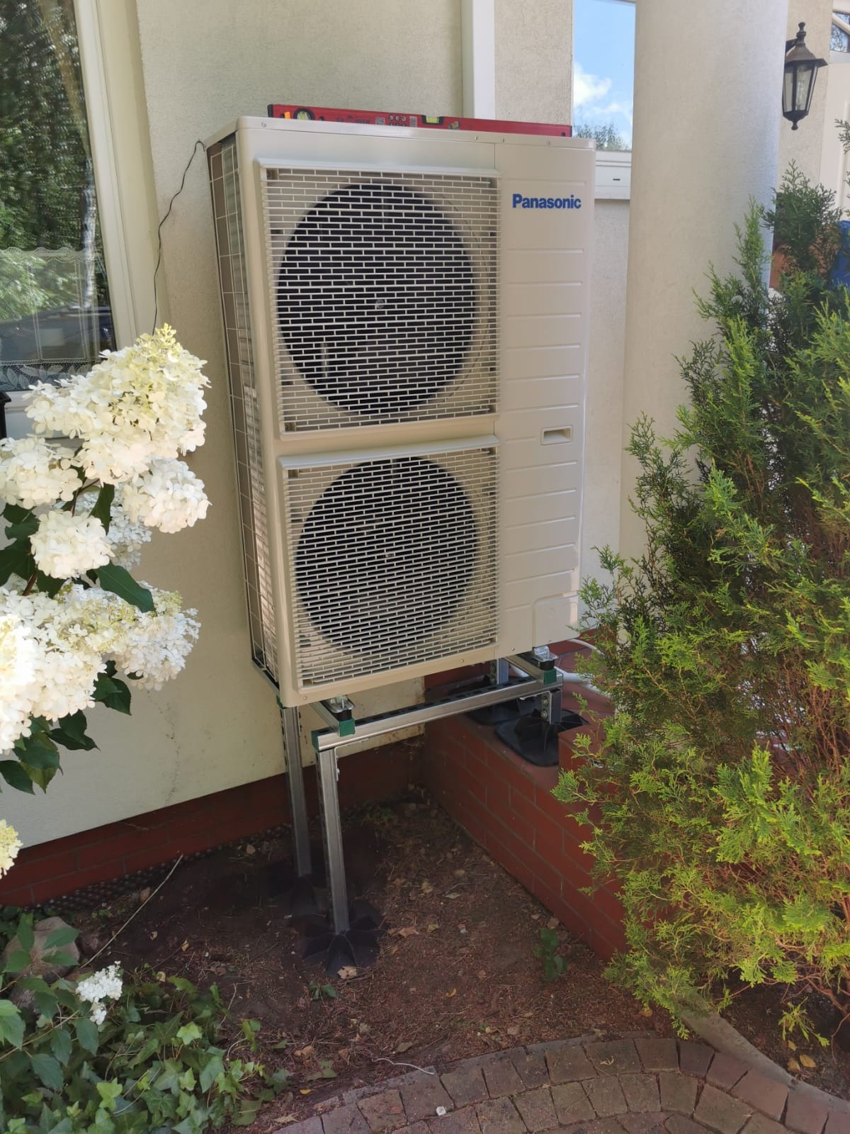 Zewnętrzna jednostka klimatyzacji Panasonic zamontowana na metalowej konstrukcji przy ścianie domu, obok kwitnącej hortensji i krzewu iglastego.