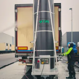 Automatyczna myjka do samochodów ciężarowych w trakcie pracy, operator w odzieży roboczej kontroluje proces, widoczny spływający strumień wody po karoserii pojazdu.