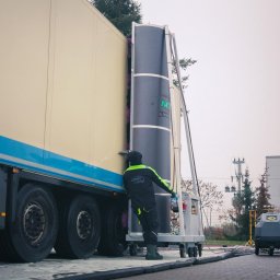 MTW Global - Pracownik w uniformie obsługuje automatyczną myjnię dla samochodów ciężarowych, czyszcząc bok naczepy; widoczne koła, logo firmy na kombinezonie i maszyna myjąca.