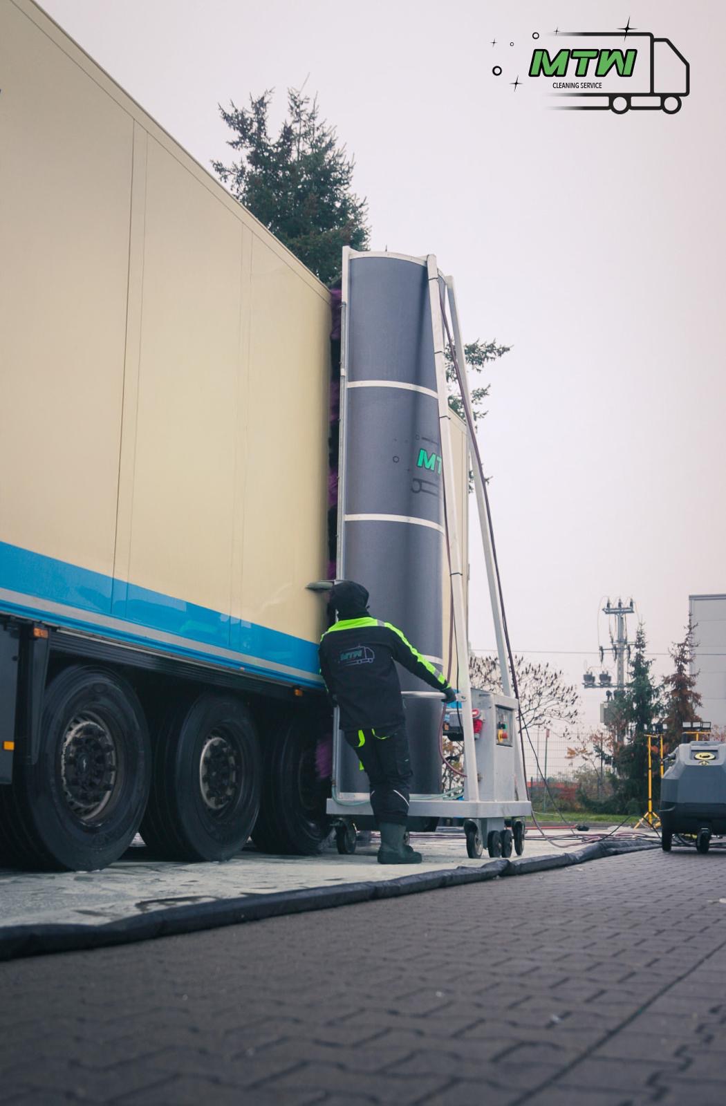 Pracownik w uniformie obsługuje automatyczną myjnię dla samochodów ciężarowych, czyszcząc bok naczepy; widoczne koła, logo firmy na kombinezonie i maszyna myjąca.