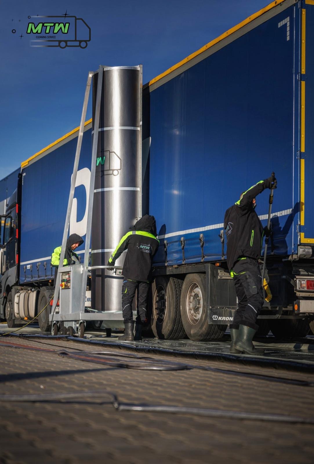 Dwóch pracowników w kombinezonach firmowych czyści bok niebieskiej naczepy ciężarówki za pomocą mobilnej platformy roboczej z wysięgnikiem i szczotki na długim kiju, na jasnym brukowanym placu.