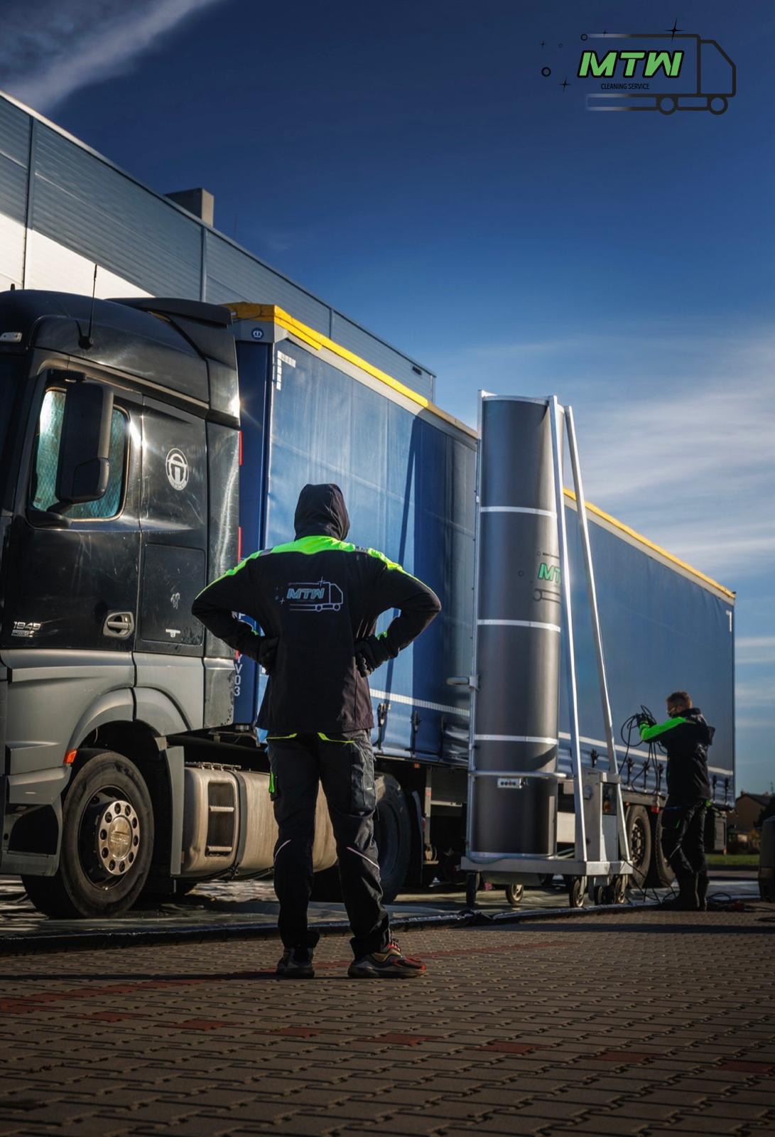 Pracownik w firmowej kurtce z logo MTW obserwuje proces czyszczenia naczepy tira za pomocą specjalistycznego urządzenia na kółkach, drugi pracownik obsługuje urządzenie.