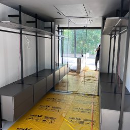 Profi Meble Oleksii Sainchyn - Nowoczesna garderoba z szarymi szafkami i czarnymi elementami konstrukcyjnymi, zabezpieczona żółtą folią ochronną na podłodze. Widoczne lustrzane drzwi i osoba w tle.
