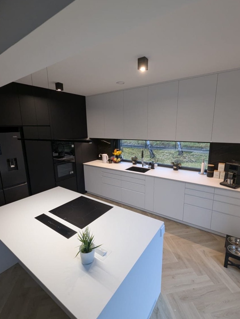 Nowoczesna kuchnia z wyspą i minimalistycznymi frontami w bieli i czerni. Parkiet w jodełkę, okno nad blatem. Roślina w doniczce na wyspie.