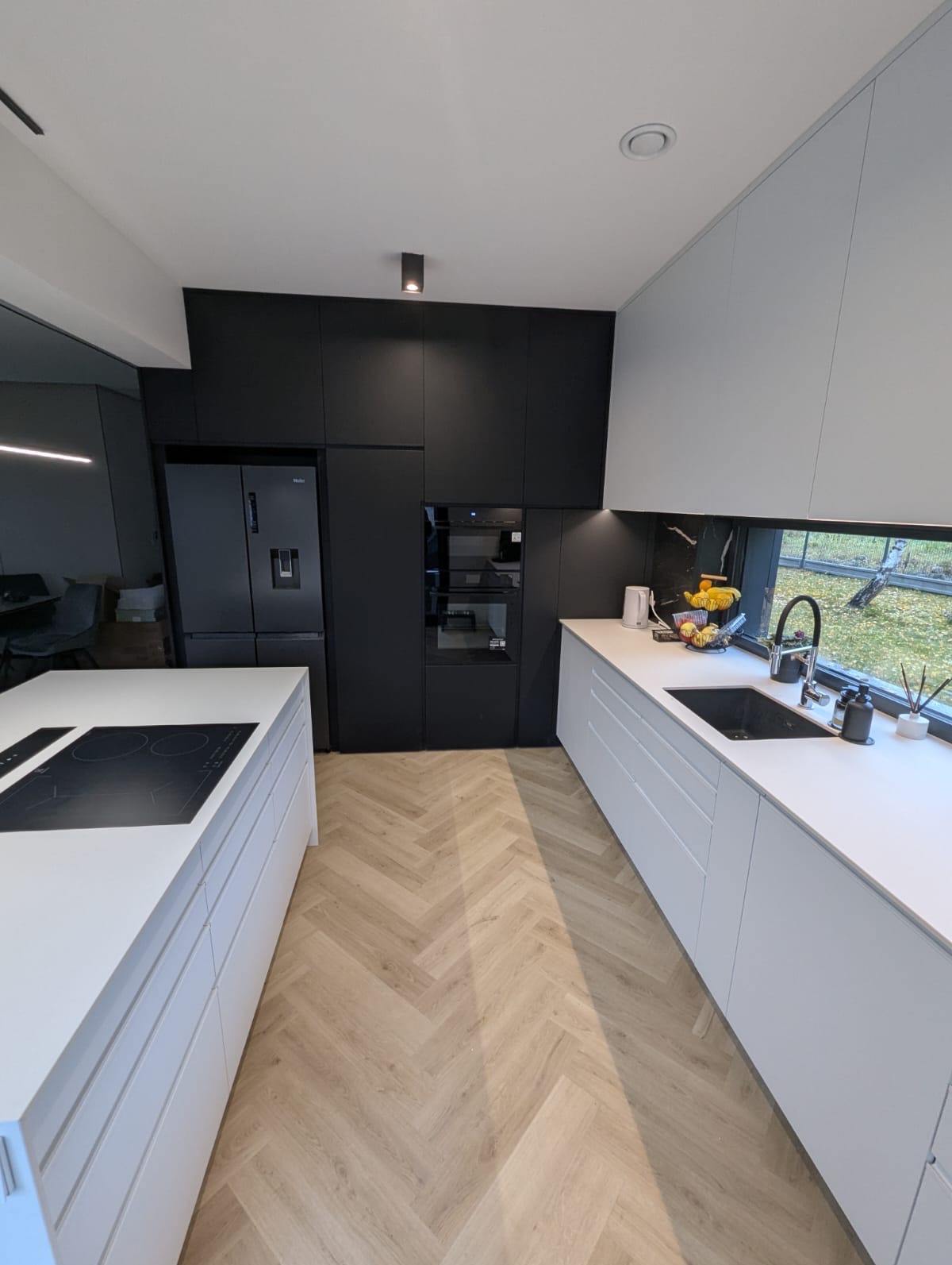 Nowoczesna kuchnia z białymi szafkami i wyspą, czarne szafki wiszące, parkiet w jodełkę, okno z widokiem na zieleń. Minimalistyczny design, oświetlenie punktowe.