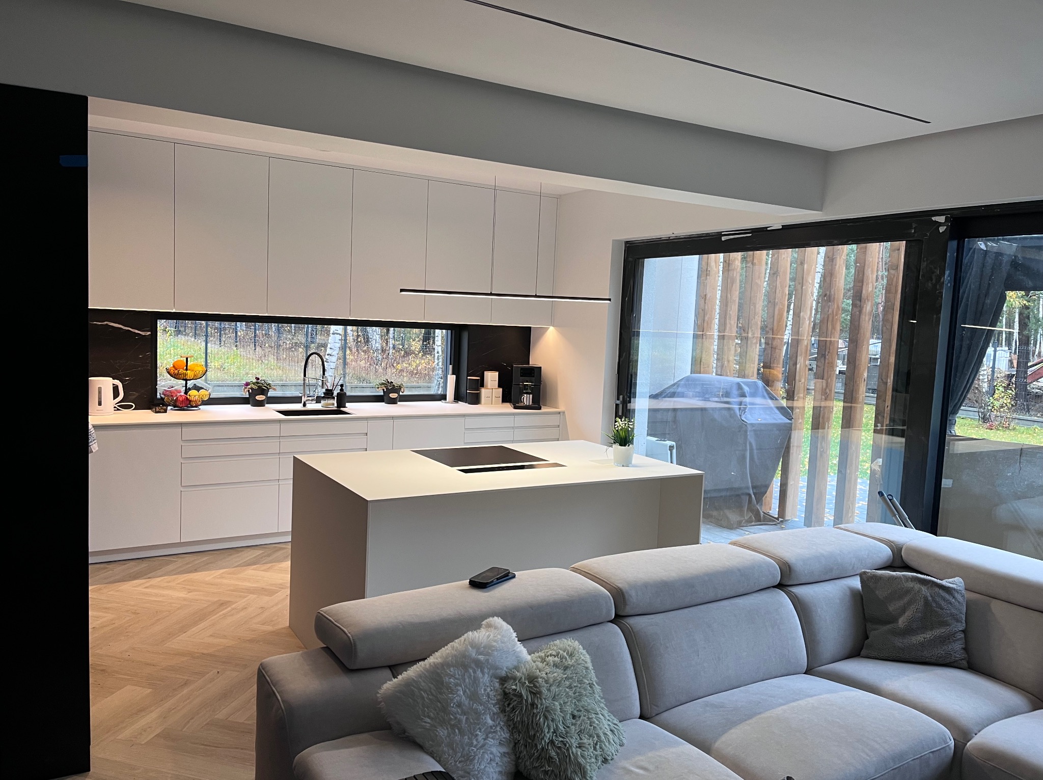 Nowoczesna, minimalistyczna kuchnia z wyspą i białymi frontami, widok z salonu. Parkiet w jodełkę, duże okno z widokiem na zewnątrz, szara sofa z poduszkami.