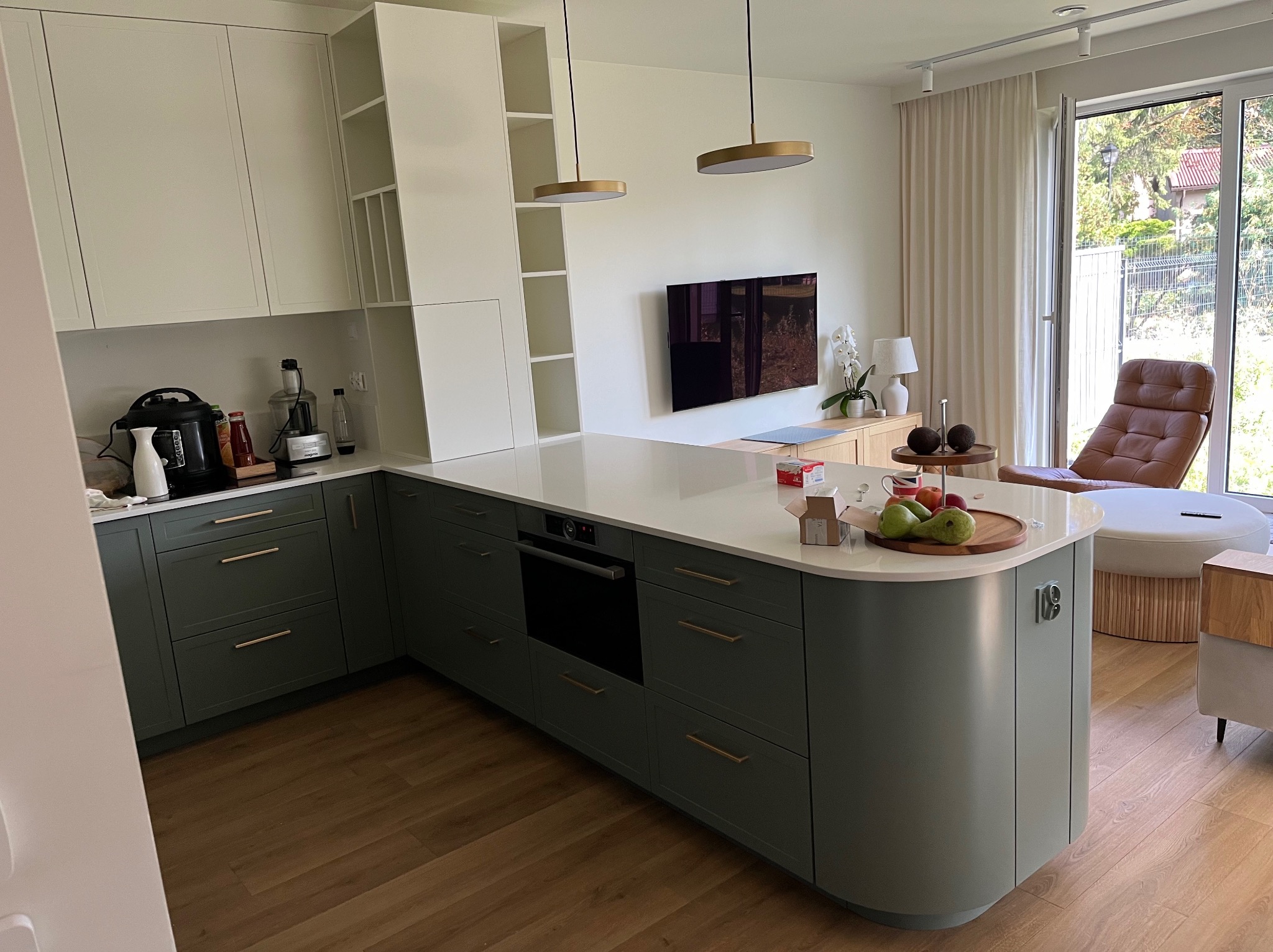 Nowoczesna kuchnia z wyspą w odcieniach zieleni i bieli, z elementami drewna i złotymi lampami wiszącymi. Widok na salon z brązowym fotelem i dużym oknem.