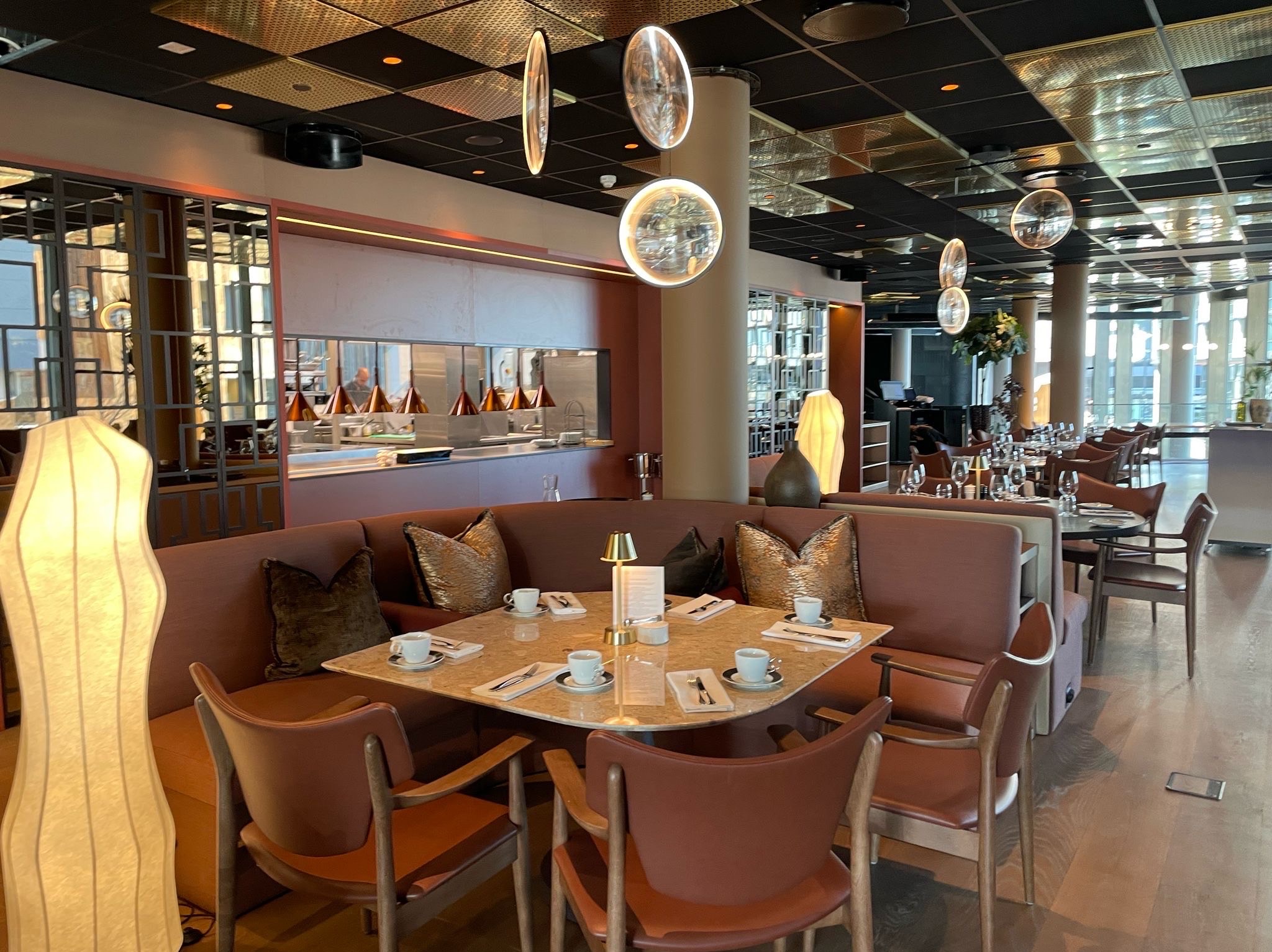 Elegancka aranżacja wnętrza restauracji z marmurowym stołem, brązowymi krzesłami i sofą. Dekoracyjne lampy wiszące i złote akcenty na suficie dodają luksusowego charakteru.