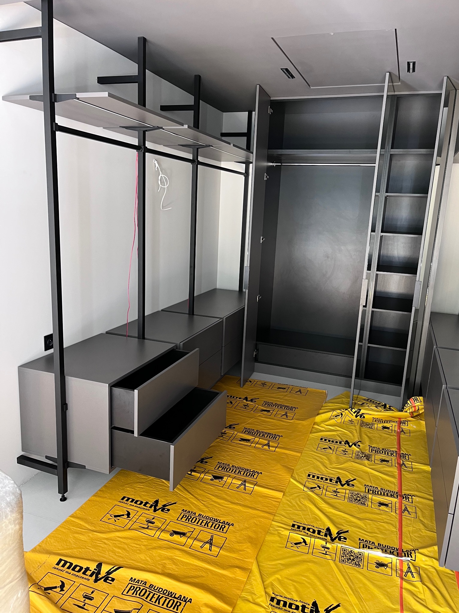 Nowoczesna garderoba w szarościach z otwartymi półkami i szafą z drzwiami, zabezpieczona folią ochronną na podłodze. Widoczne detale wykończenia i minimalistyczny design.