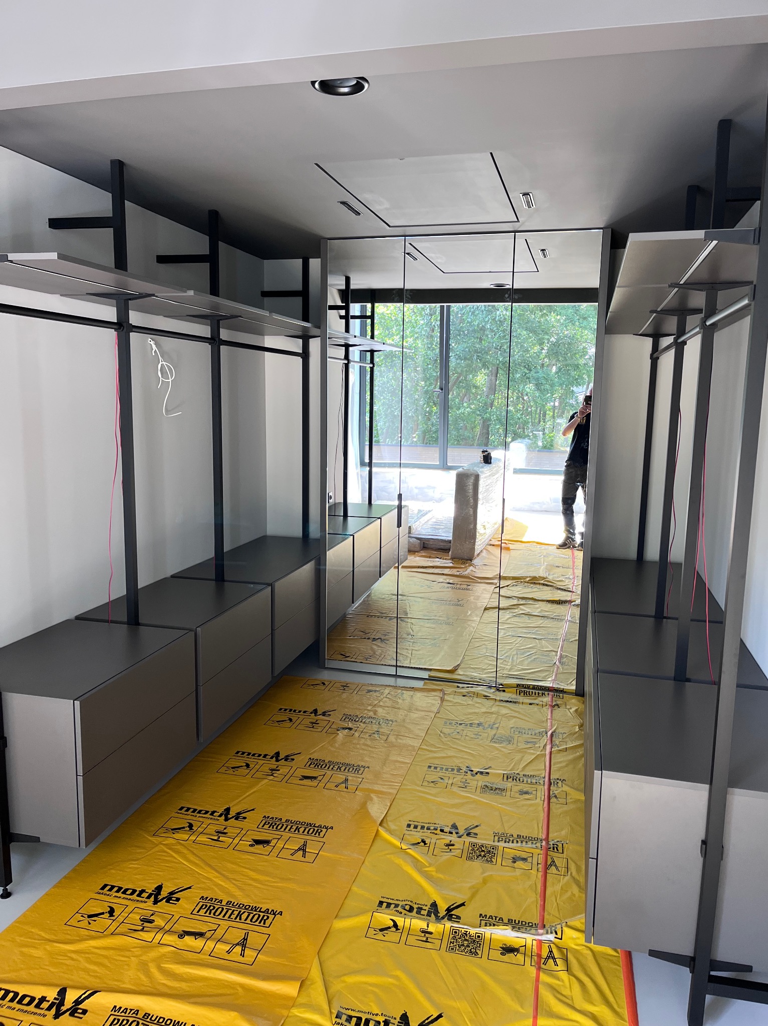 Nowoczesna garderoba z szarymi szafkami i czarnymi elementami konstrukcyjnymi, zabezpieczona żółtą folią ochronną na podłodze. Widoczne lustrzane drzwi i osoba w tle.