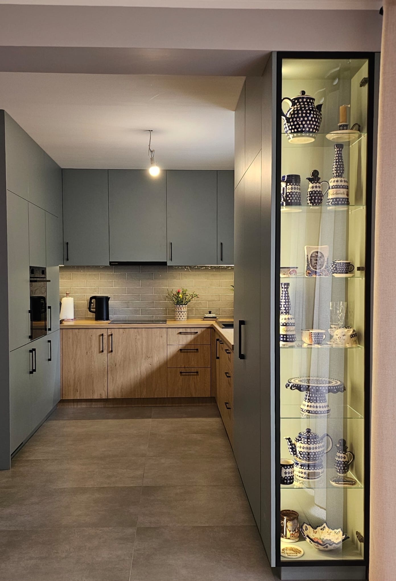 Nowoczesna kuchnia z szarymi i drewnianymi szafkami, oświetleniem podszafkowym i przeszkloną witryną z kolekcją ceramiki w niebieskie wzory.