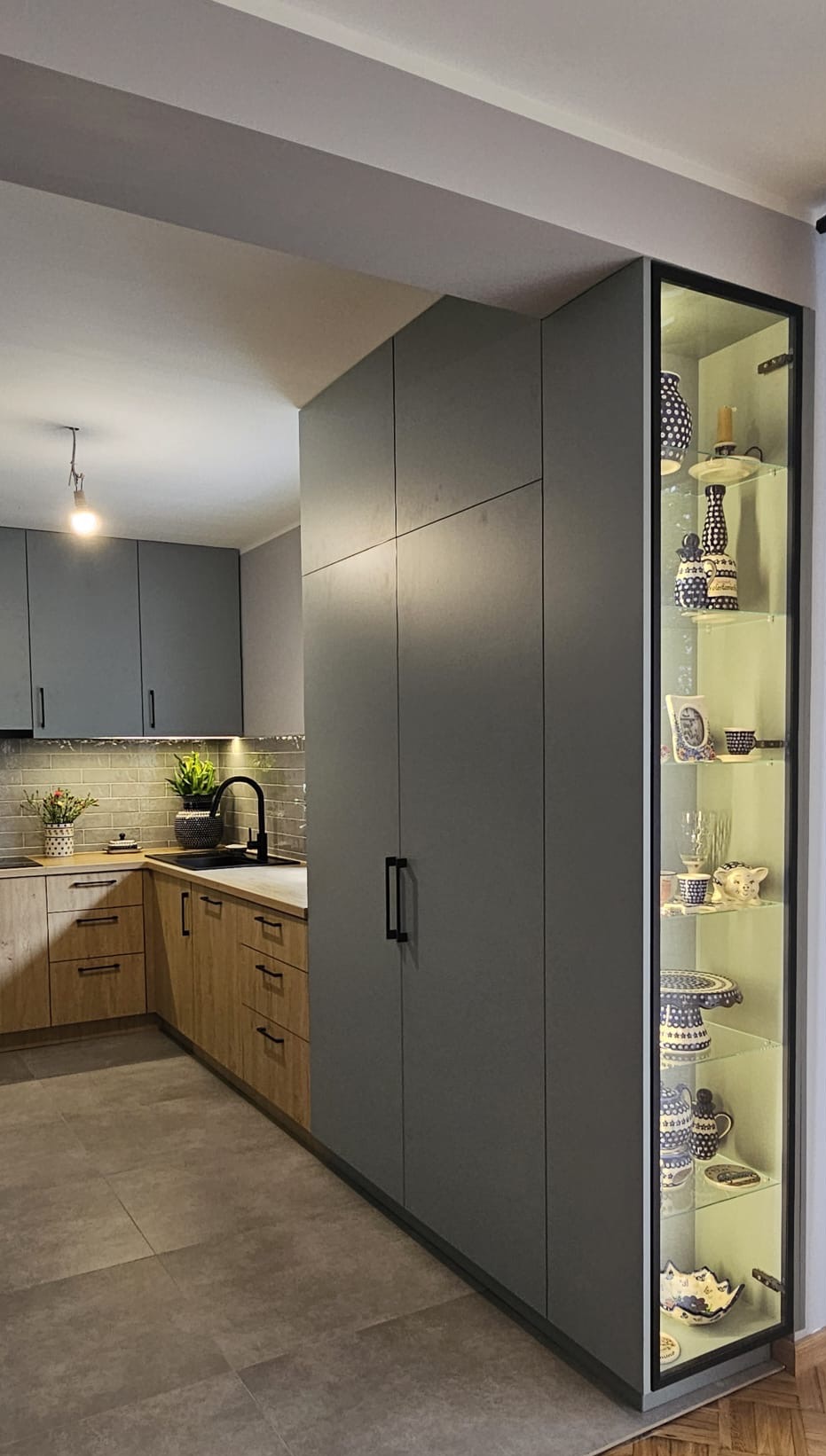 Nowoczesna kuchnia z szarymi szafkami, drewnianymi frontami i przeszkloną witryną z oświetleniem, eksponującą kolekcję ceramiki w niebieskie wzory.