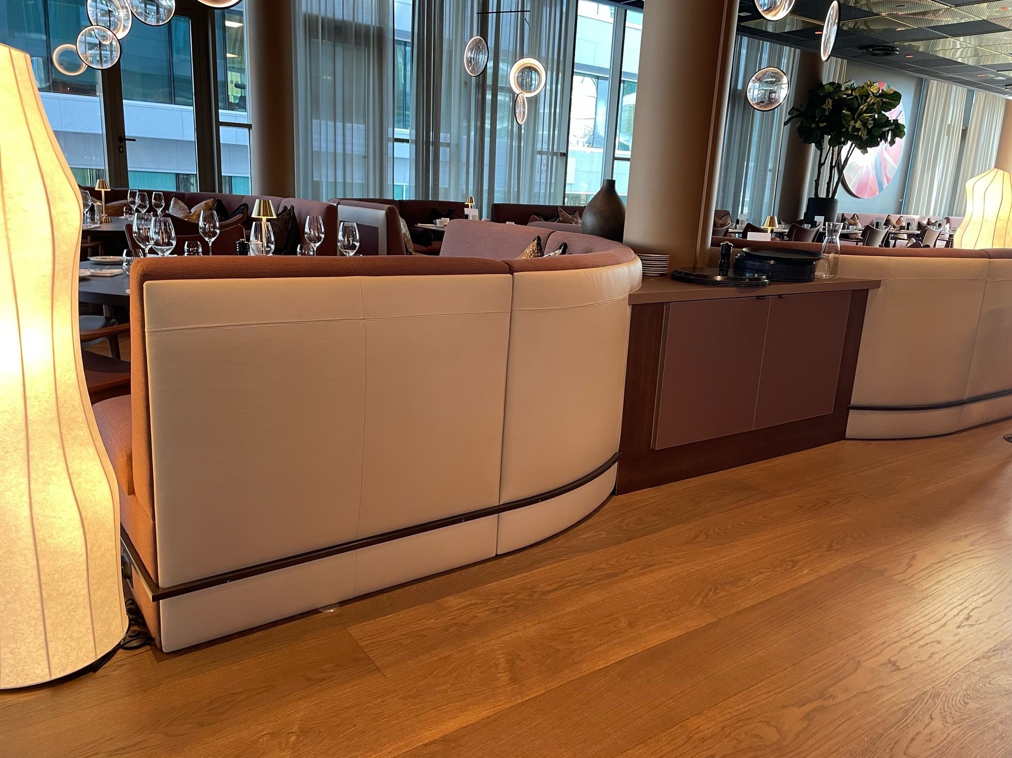 Eleganckie, zaokrąglone sofy w restauracji z widokiem na miasto. Jasne obicia, drewniane wykończenia i parkiet tworzą przytulną atmosferę. Stoliki nakryte do kolacji.