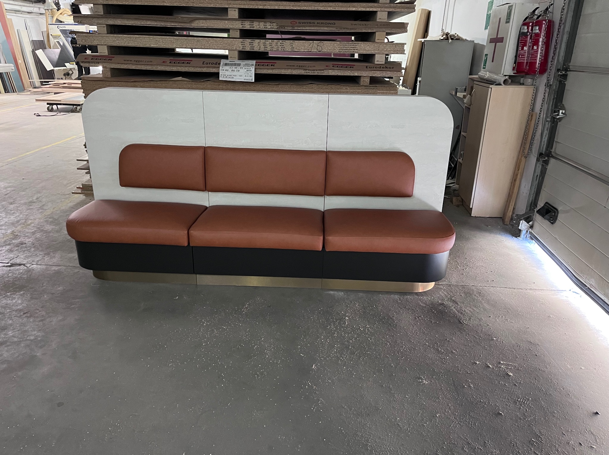 Tapicerowana ławka z oparciem w pracowni meblarskiej, na tle płyt meblowych. Widoczne detale wykończenia i materiały. Ujęcie z perspektywy podłogi.