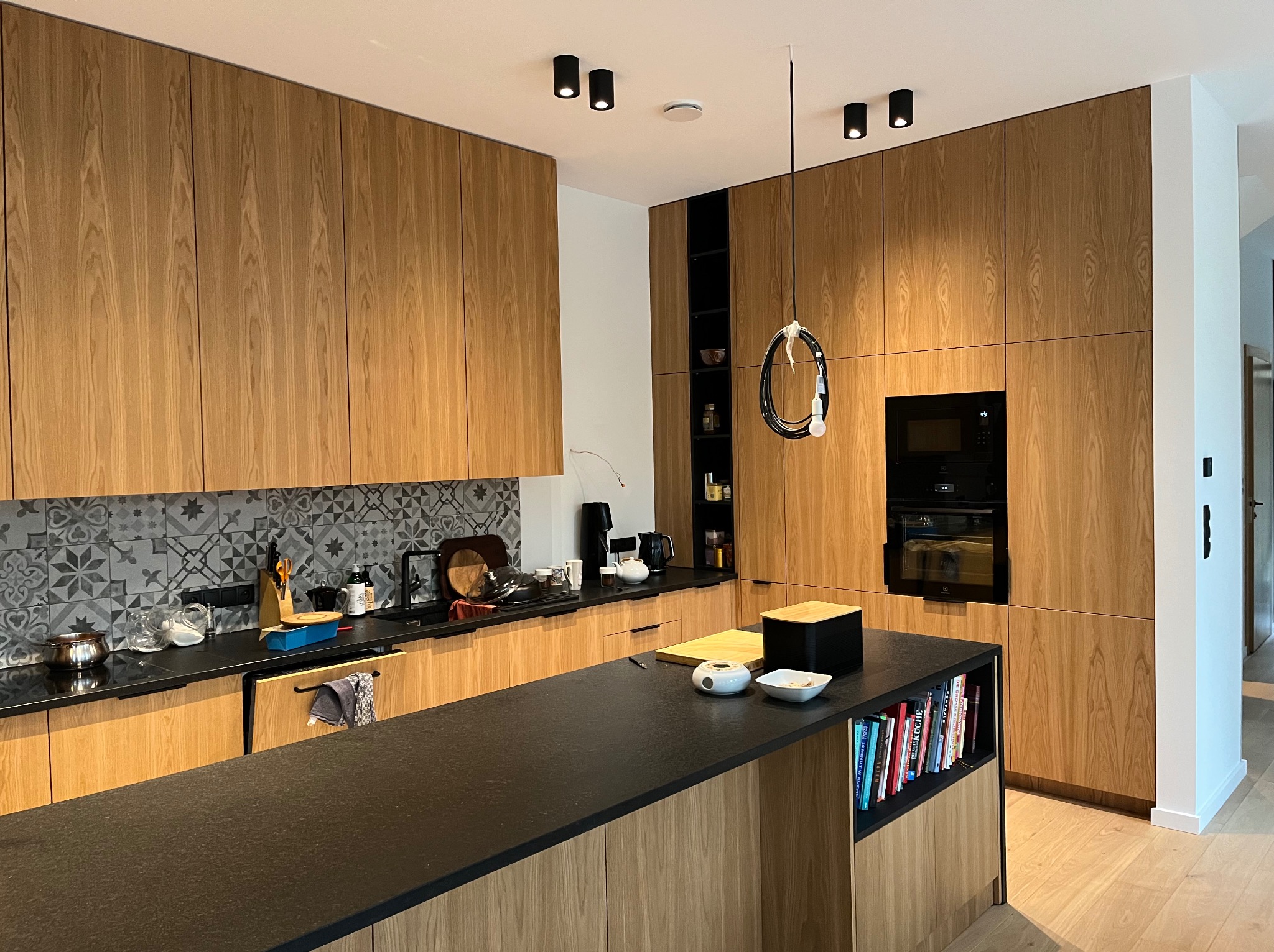Nowoczesna kuchnia z drewnianymi frontami, czarnym blatem i wyspą. Dekoracyjne płytki nad blatem, minimalistyczne oświetlenie sufitowe i wisząca lampa nad wyspą.