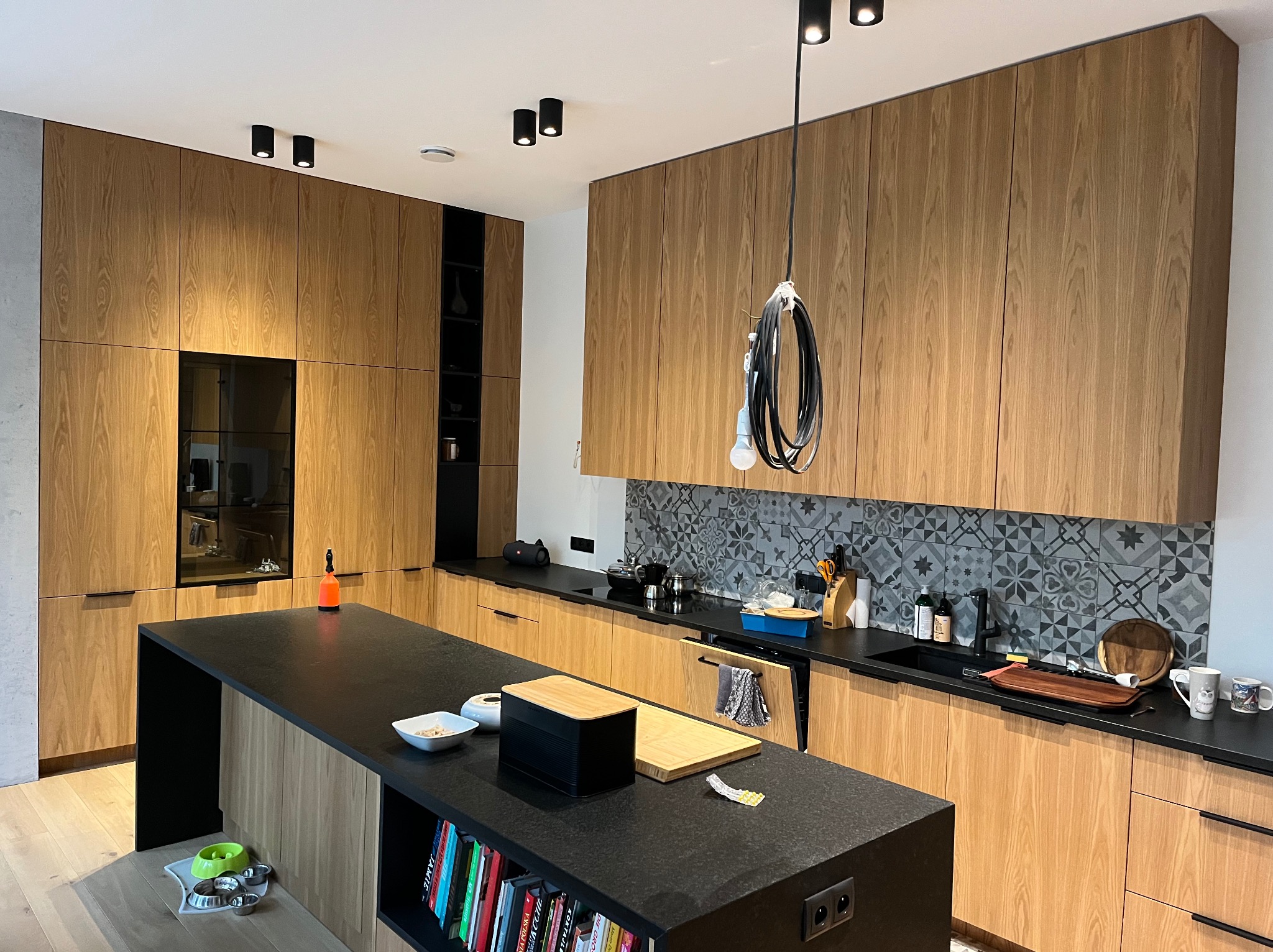 Nowoczesna kuchnia z drewnianymi frontami, czarnym blatem i wyspą. Widoczne oświetlenie punktowe i dekoracyjne płytki na ścianie. Minimalistyczny design z elementami wyposażenia.