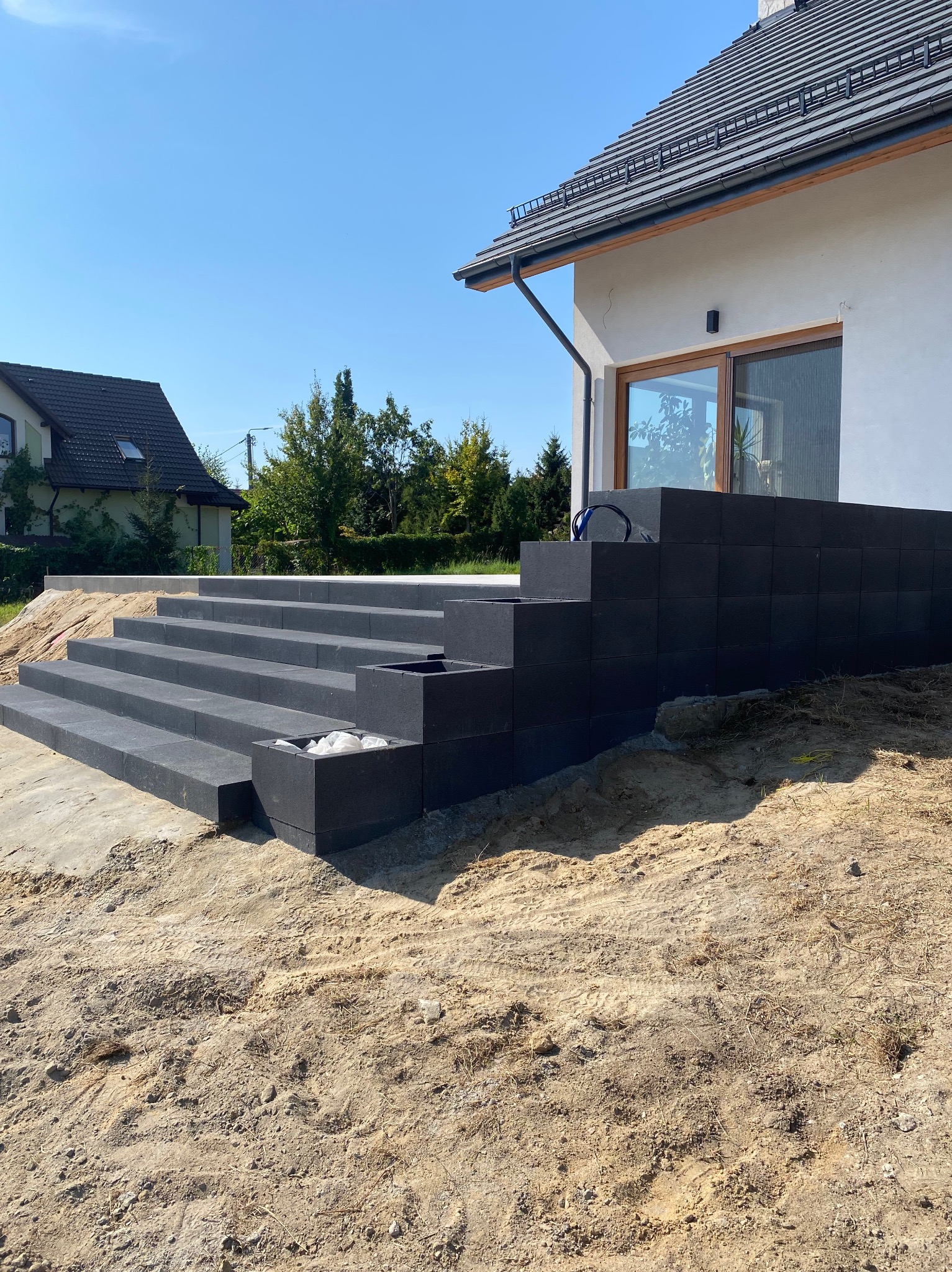 Schody z ciemnych, kwadratowych elementów prowadzące do domu z jasną elewacją, widoczne elementy krajobrazu i przygotowany teren.