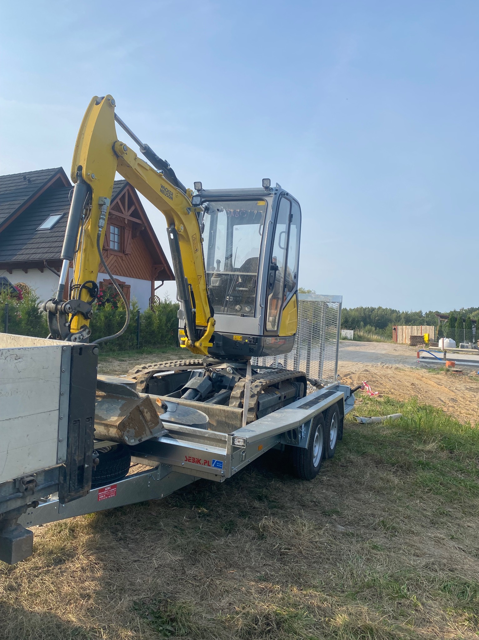 Żółto-szara mini koparka Wacker Neuson na platformie przyczepy, widok z boku, w tle dom jednorodzinny i teren budowy.