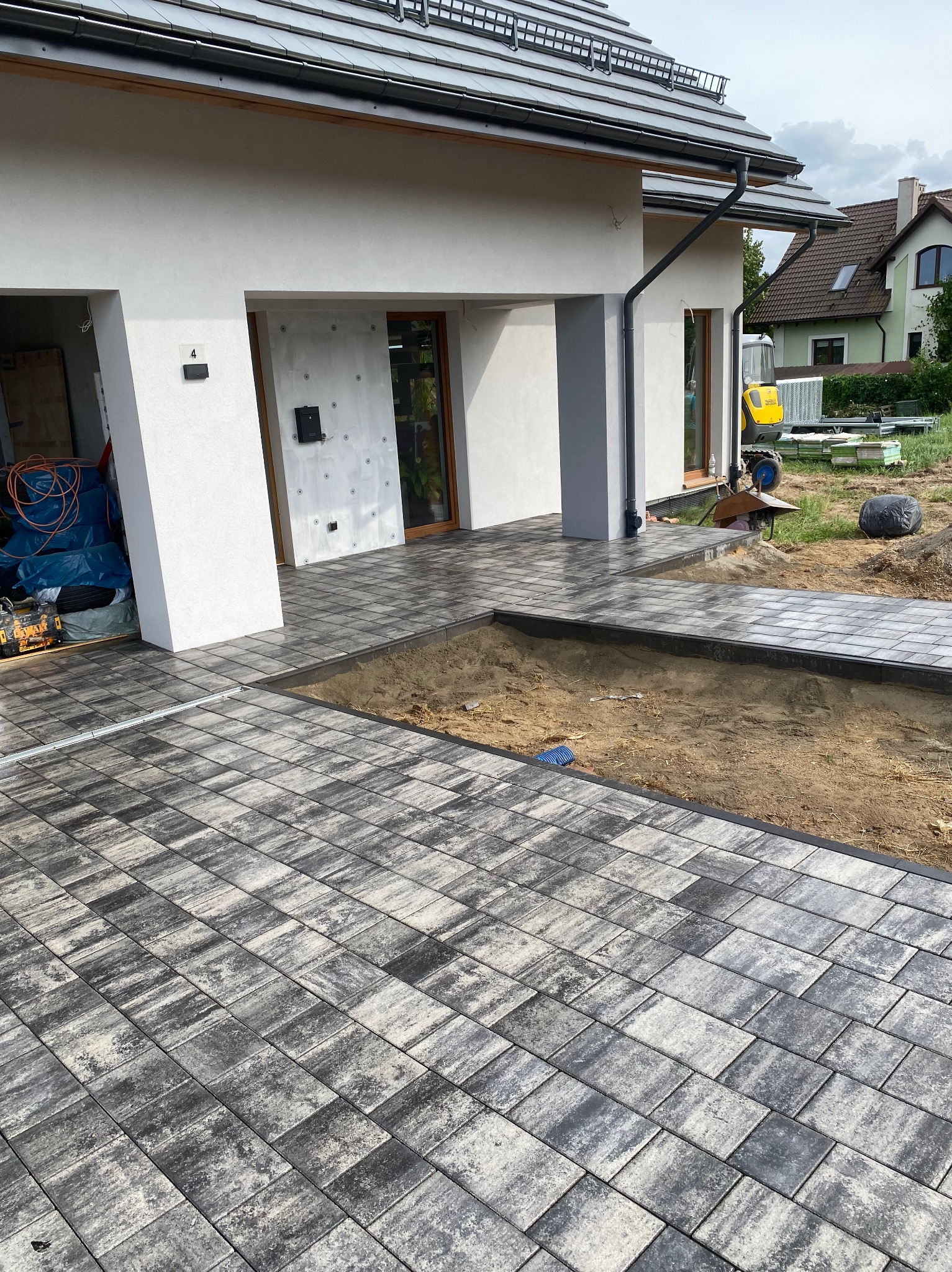 Częściowo ułożona kostka brukowa w odcieniach szarości przed nowoczesnym domem z białymi ścianami, widoczny wykop pod dalszą część nawierzchni i minikoparka w tle.