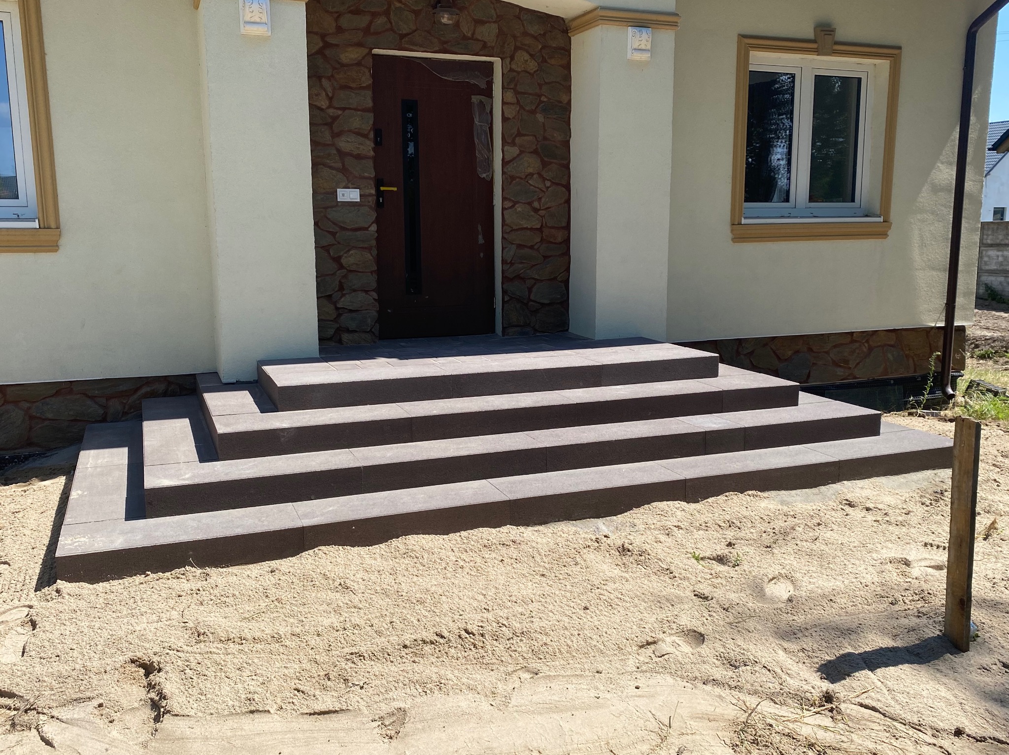 Schody wejściowe z ciemnej kostki prowadzące do drzwi wejściowych domu z elewacją z kamienia i piaskowym podłożem.