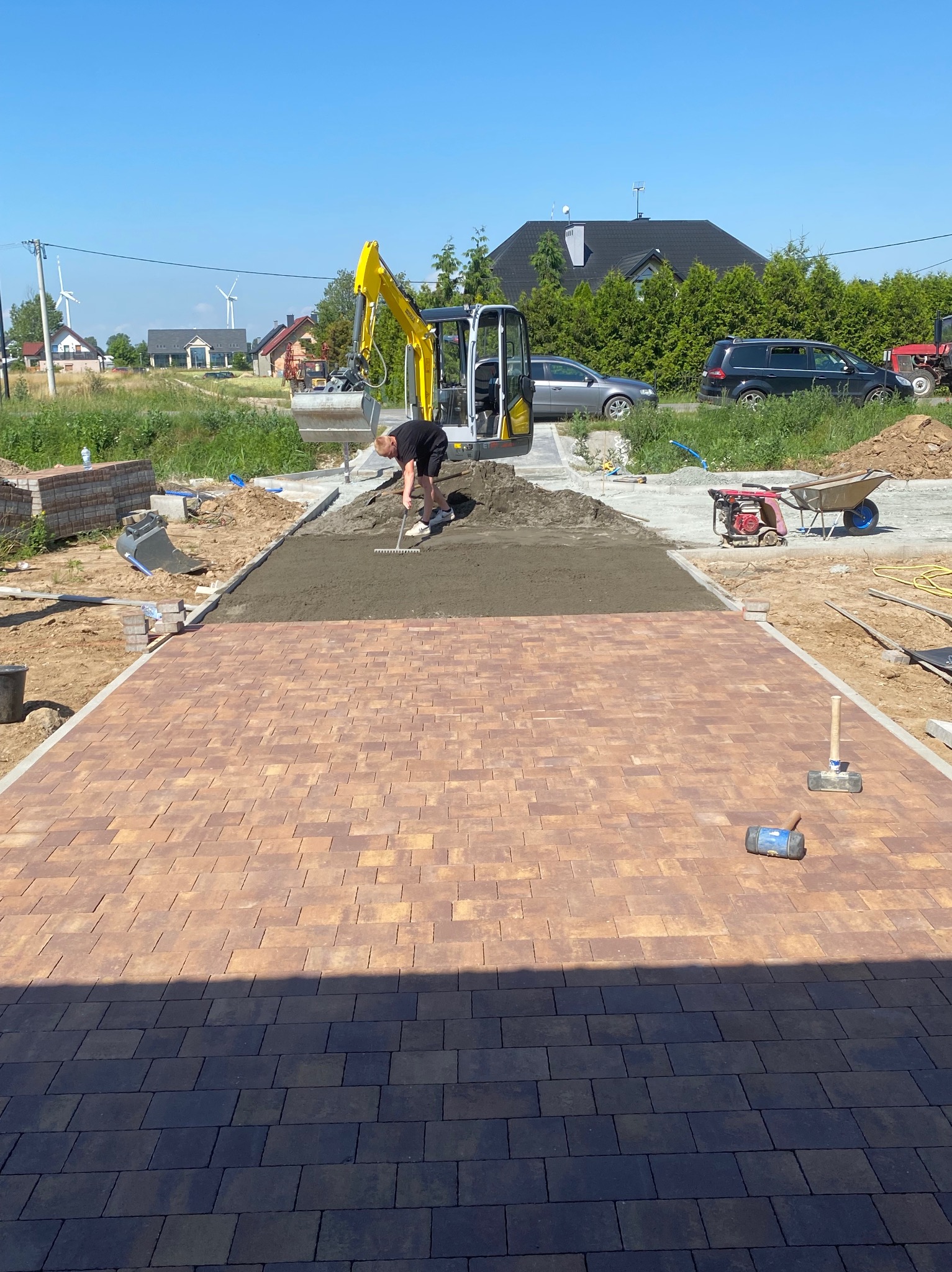 Pracownik wyrównuje piasek podłoża pod nową nawierzchnię z kostki brukowej, w tle żółta mini koparka, na obrzeżach ułożona kostka w kolorze brązowym.