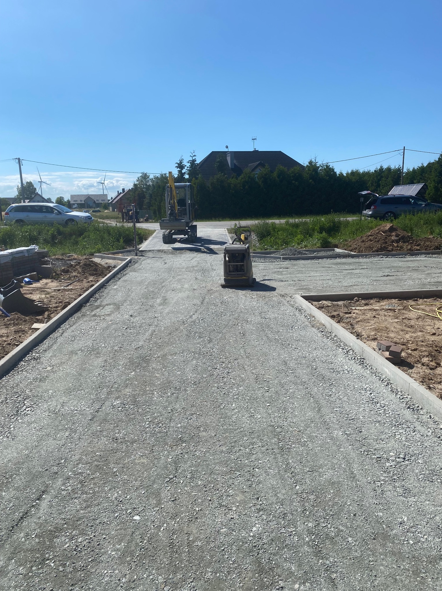 Przygotowany podkład z kruszywa pod drogę lub plac, widoczny mały walec wibracyjny i minikoparka w tle, krawężniki wyznaczają granice, słoneczny dzień.