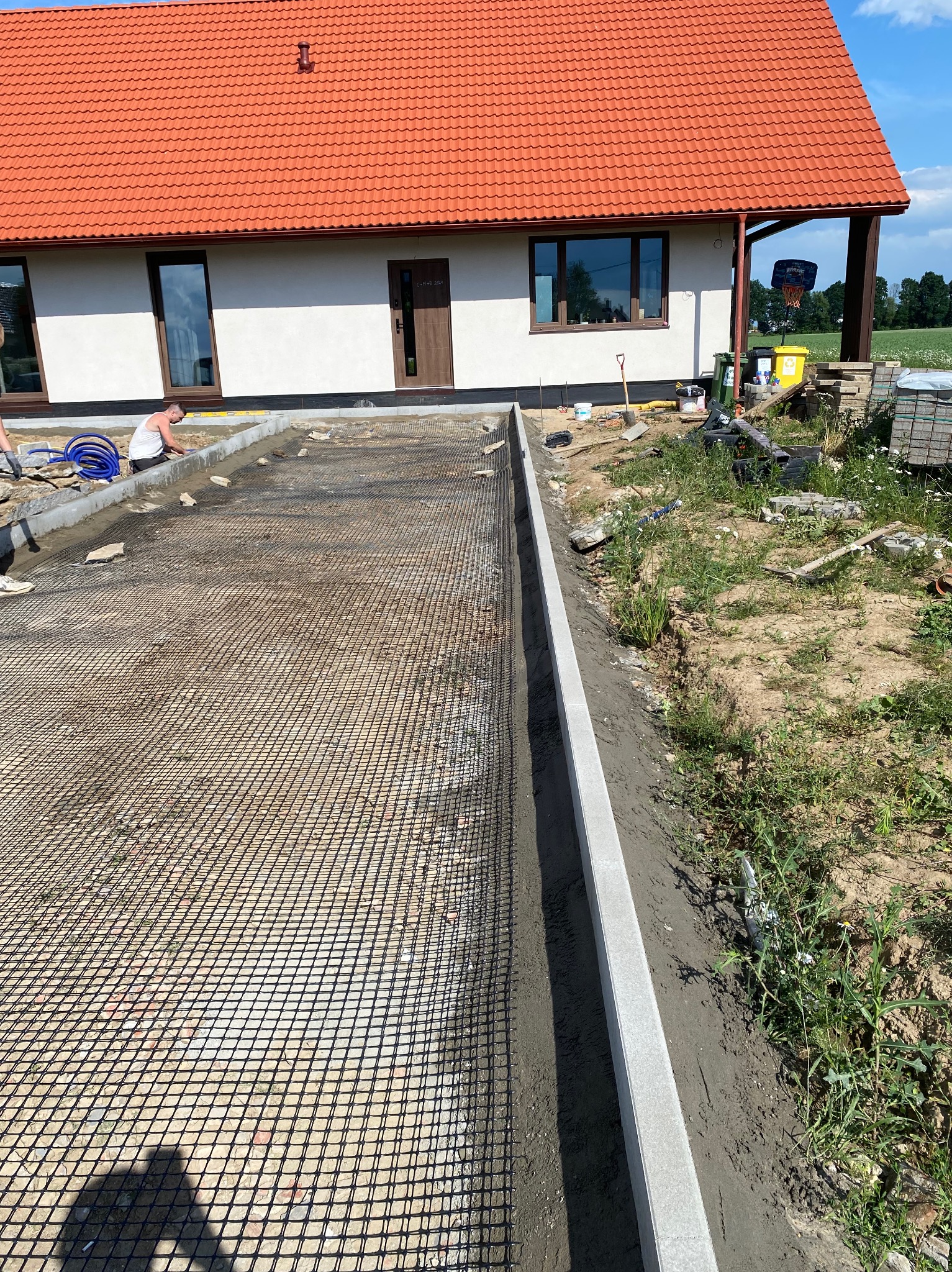 Przygotowanie podłoża pod nawierzchnię przed domem: siatka zbrojeniowa na gruncie, obrzeża betonowe w świeżym cemencie, pracownik wyrównujący krawężnik, dom z czerwoną dachówką w tle.