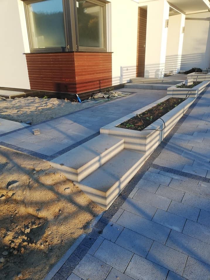 Nowoczesna posesja z szarej kostki brukowej, schody z kostki prowadzące do domu, obrzeża z ciemnej kostki, rabaty kwiatowe wzdłuż ścieżki, widoczne węże do nawadniania.