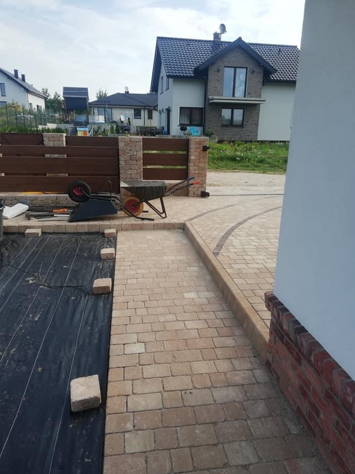 Ułożona z beżowej kostki brukowej ścieżka w ogrodzie, obok czarna agrowłóknina, w tle ogrodzenie z drewnianymi panelami i domy.