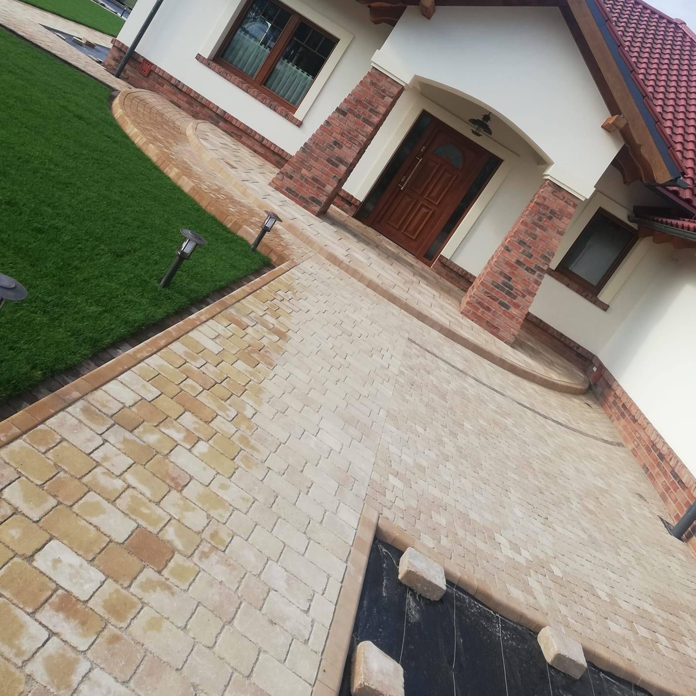 Ułożona, jasna kostka brukowa prowadząca do wejścia domu z ceglaną elewacją, widoczne lampy ogrodowe i trawnik.