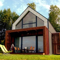 M&M projekt sp. z o.o. - Nowoczesny dom z dużym przeszkleniem szczytowym i drewnianą pergolą, zieleń w tle, dwa zielone leżaki na trawniku.